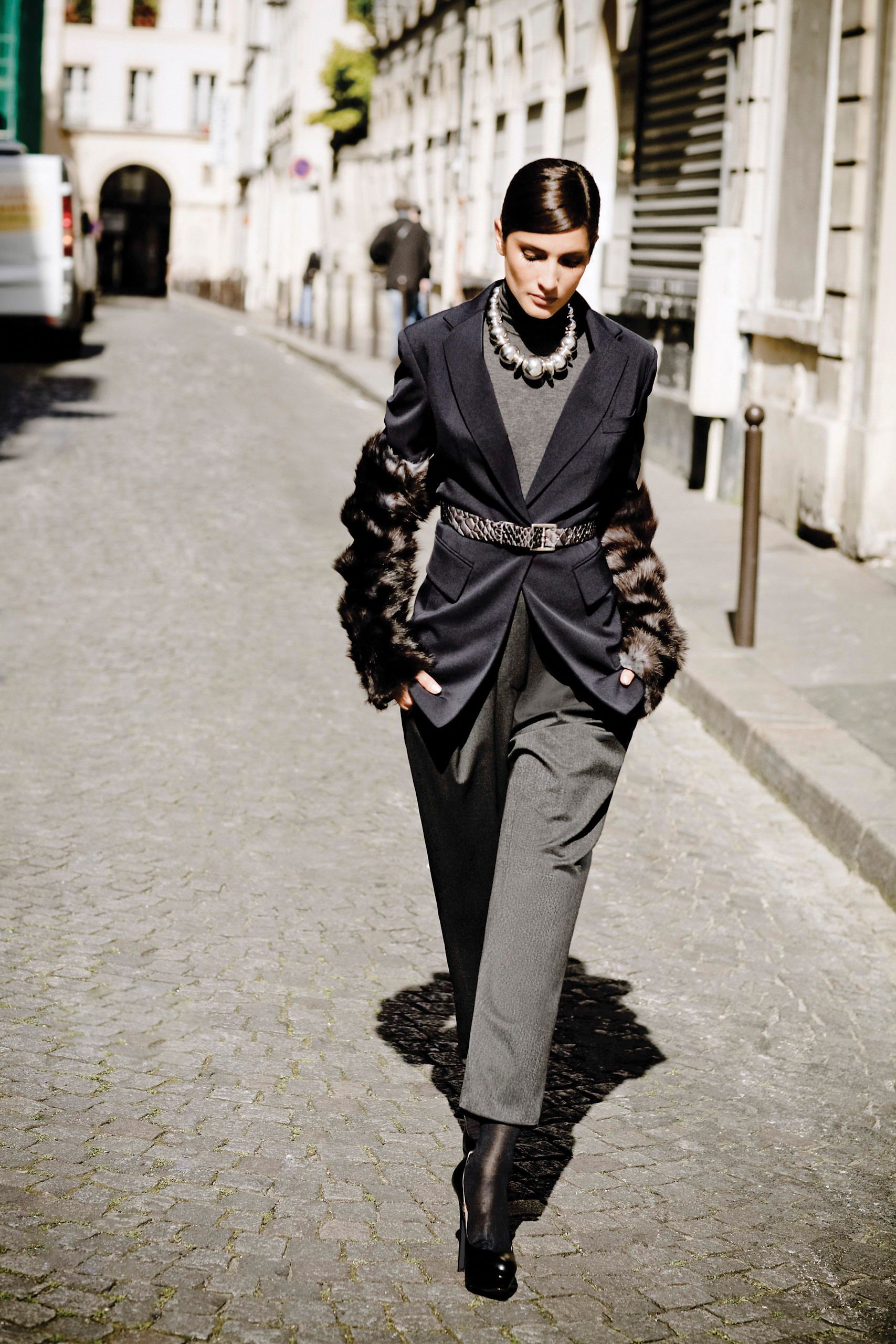A model walks a cobblestone street in a dark blazer with fur sleeves, gray trousers, belt, and statement necklace.