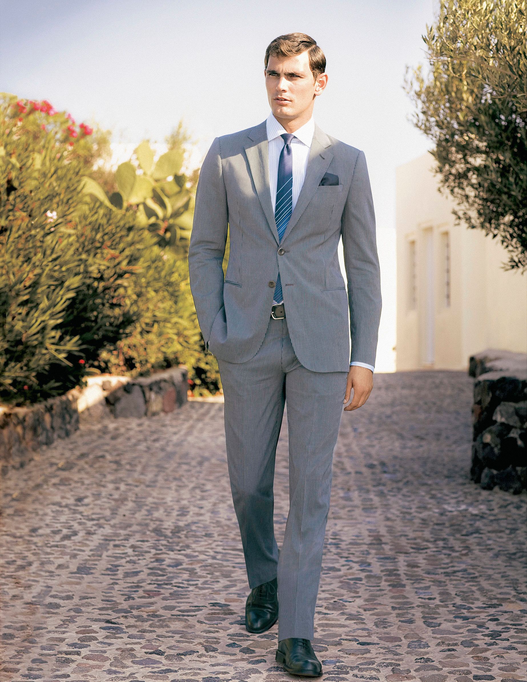 Man in a slim-fit striped suit with blue tie and pocket square, standing on a cobblestone path outdoors.