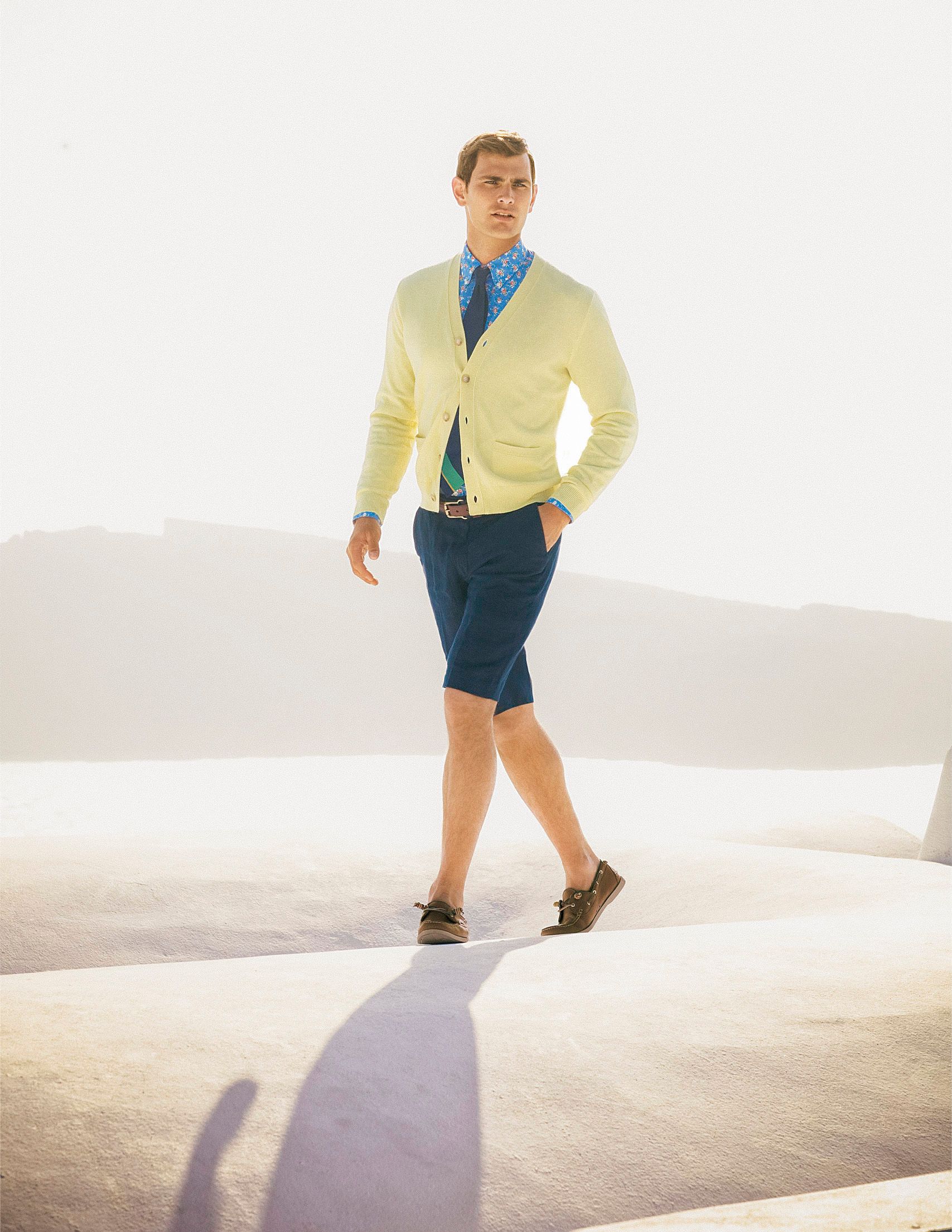Man walking in yellow cardigan, blue shorts, and loafers against a bright white Mediterranean backdrop.