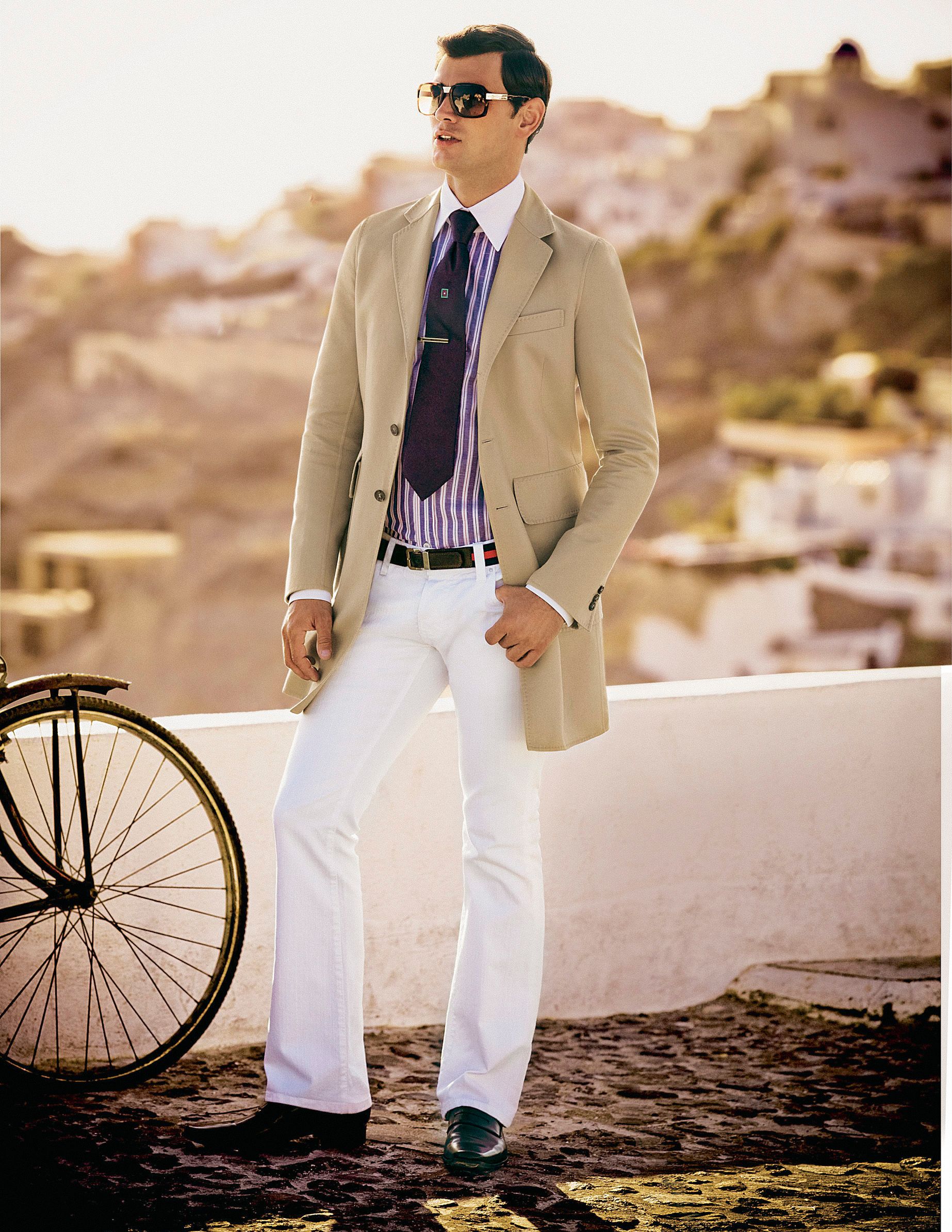 Stylish man in beige blazer, white trousers, striped tie, and sunglasses poses near a bicycle in a Mediterranean setting.