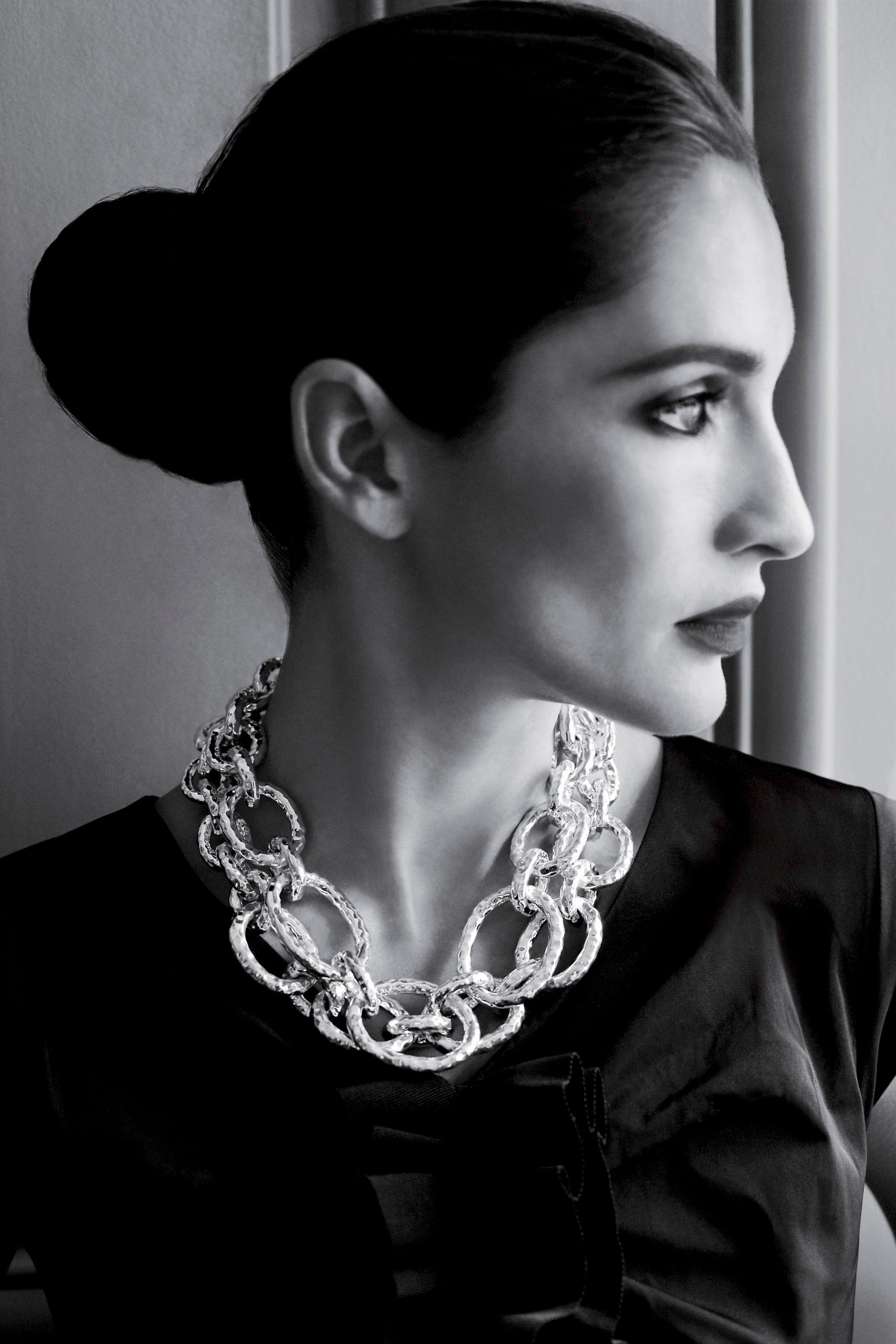 Black and white portrait of elegant woman in profile wearing bold chain-link necklace and dark blazer with hair in bun.
