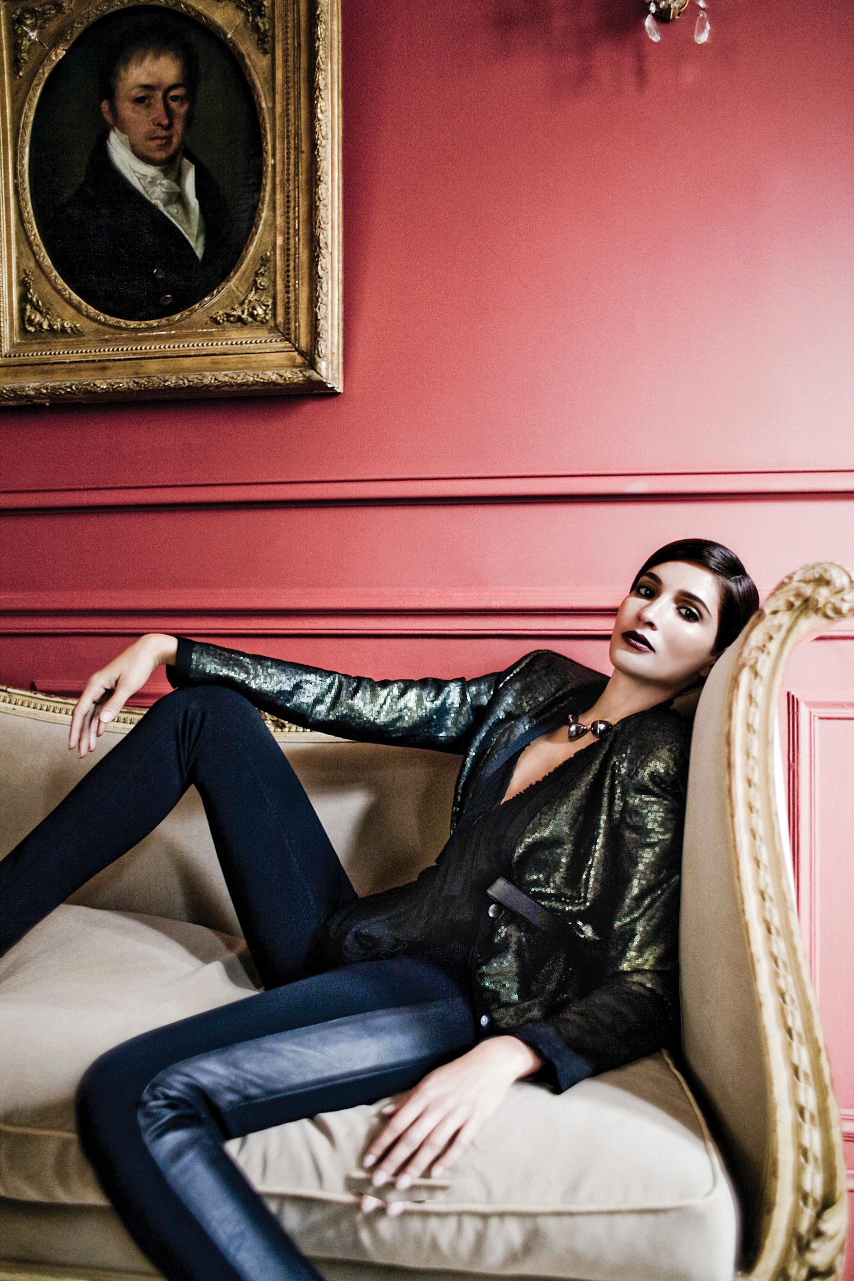 A woman in a black leather jacket and blue pants reclines on a cream sofa beneath a classic portrait on a pink wall.