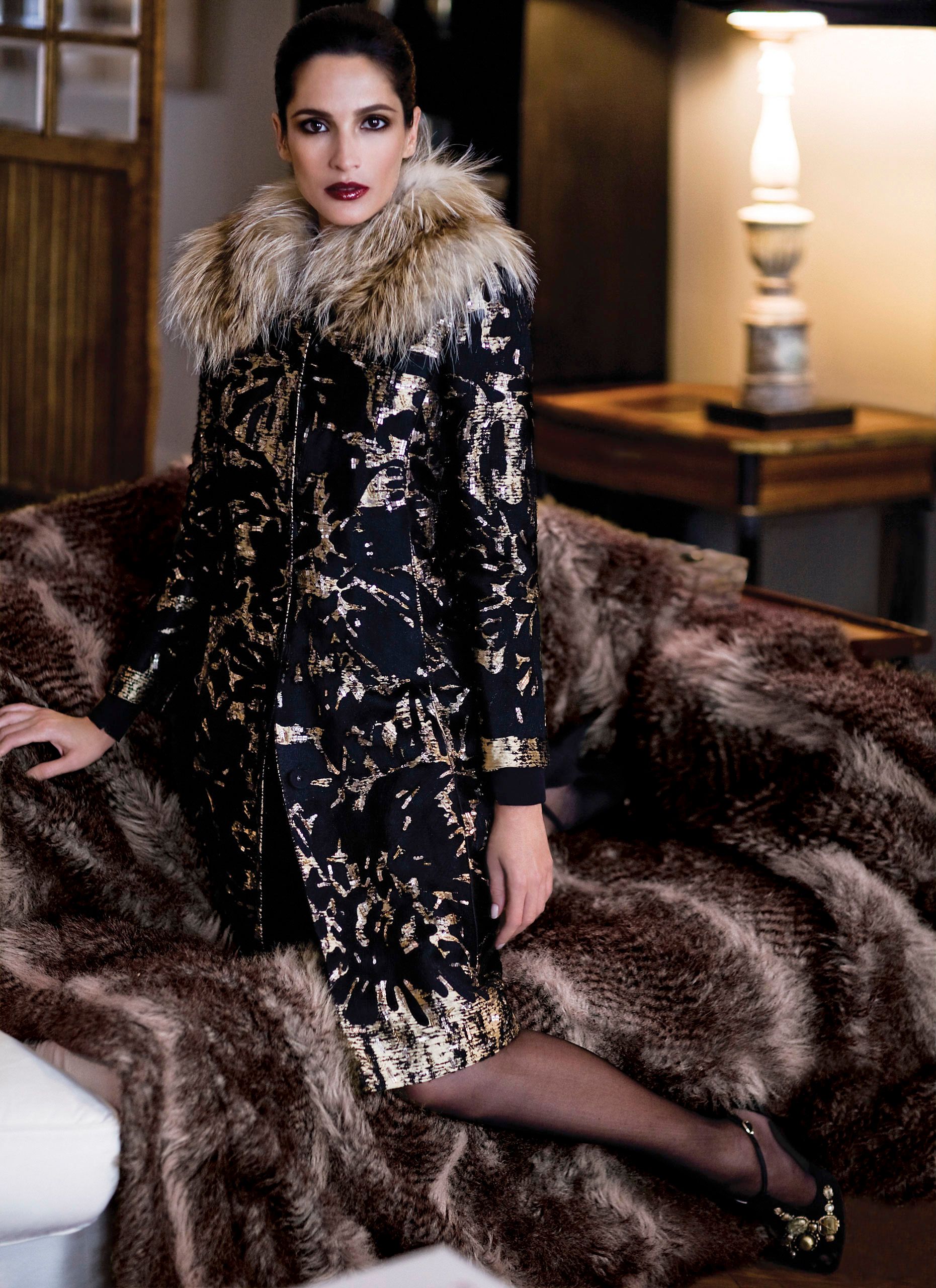 Woman in black floral brocade coat with fur collar poses on fur-draped sofa in elegant interior setting.