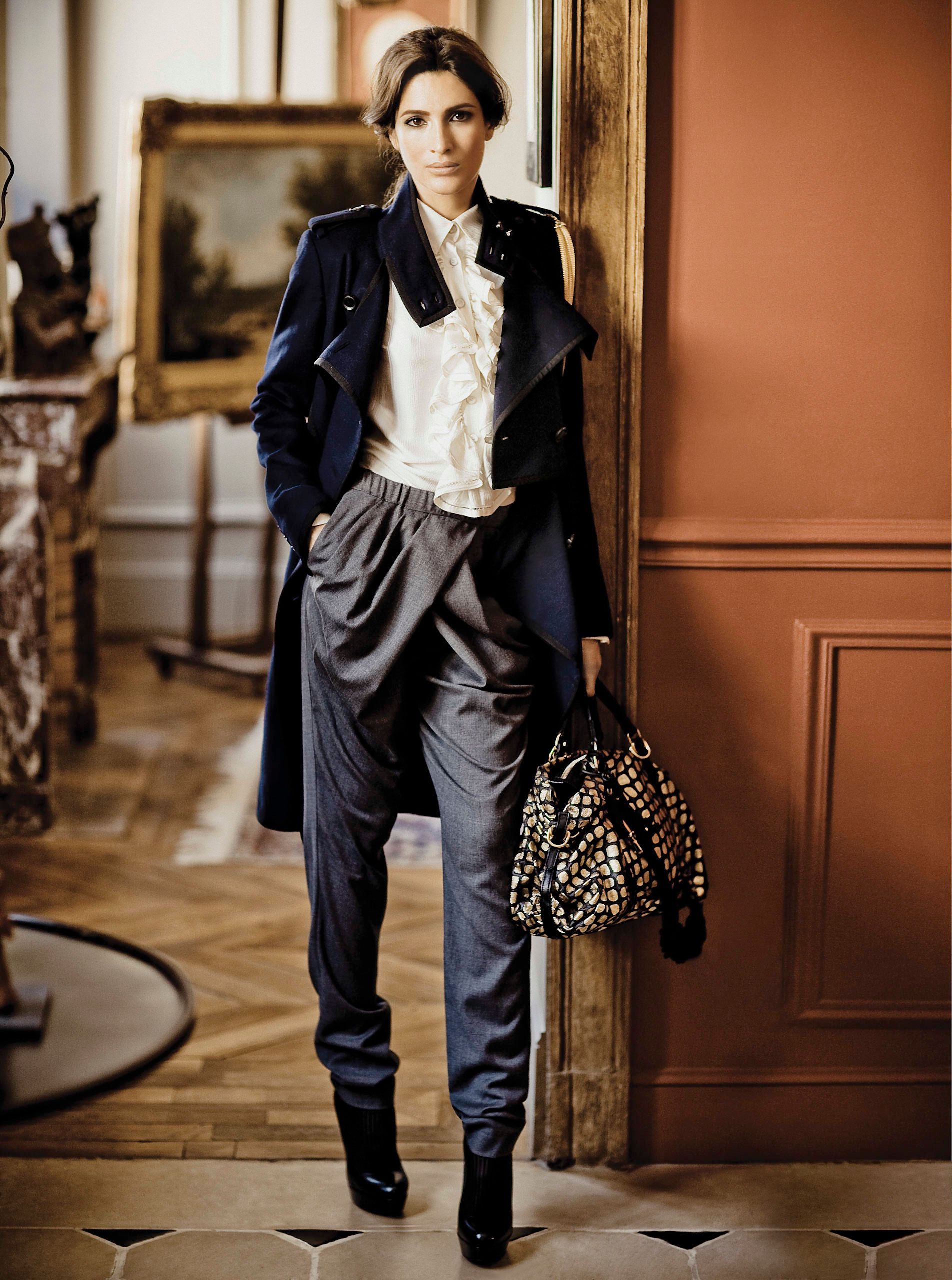Woman in navy coat, white ruffled blouse, gray trousers, and black boots holding a studded bag in an elegant room.
