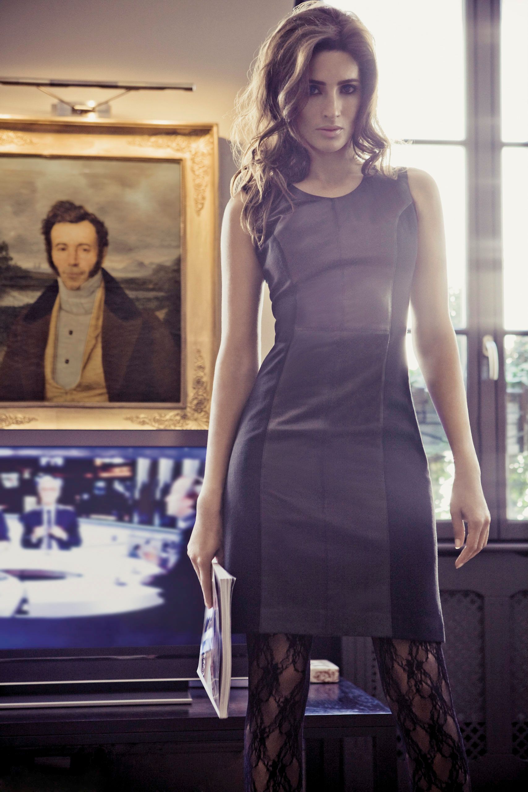 Woman in gray sheath dress and lace tights holds papers near vintage portrait painting and window.