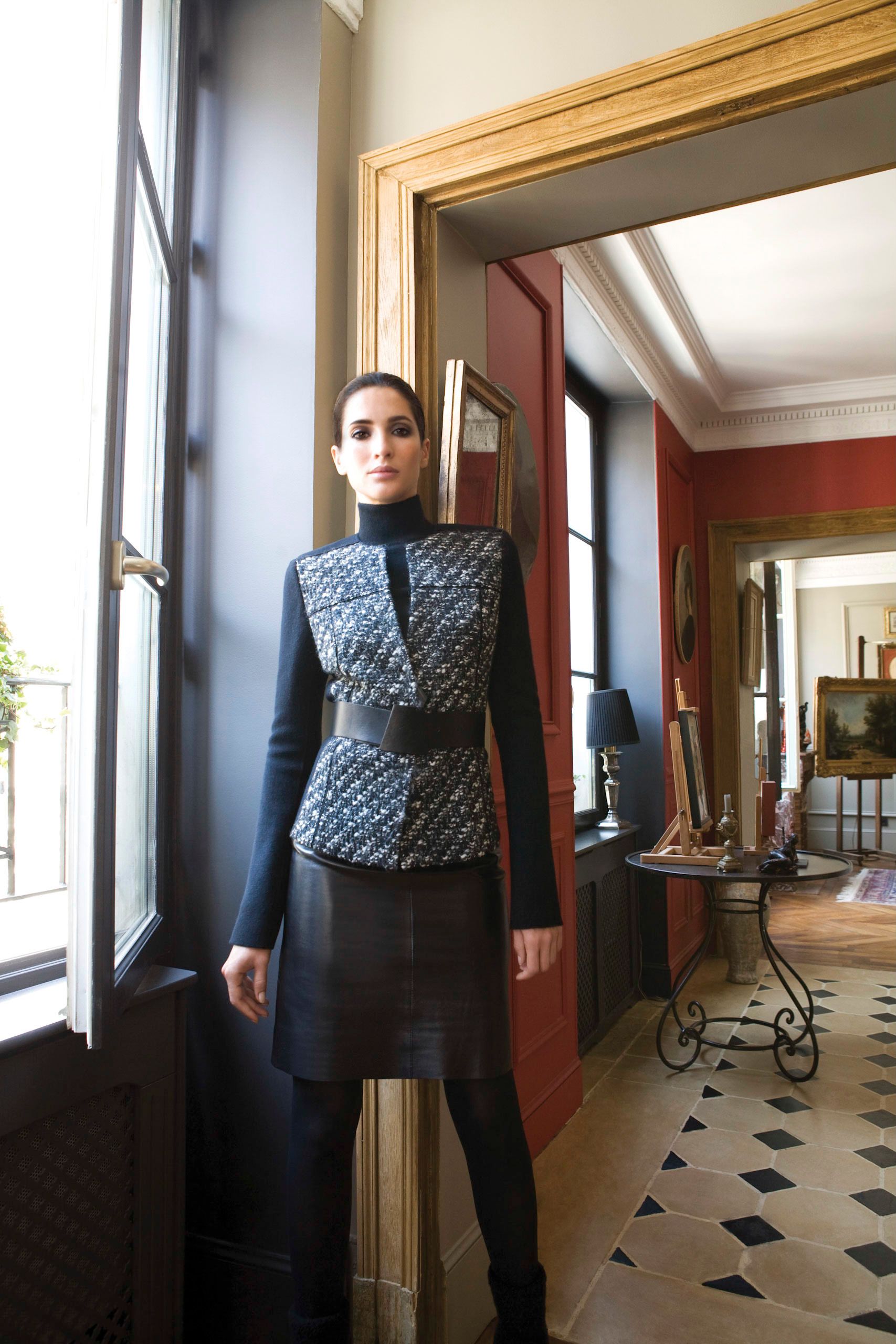 Model wearing a navy tweed structured jacket with leather skirt and wide belt in an elegant Parisian interior.