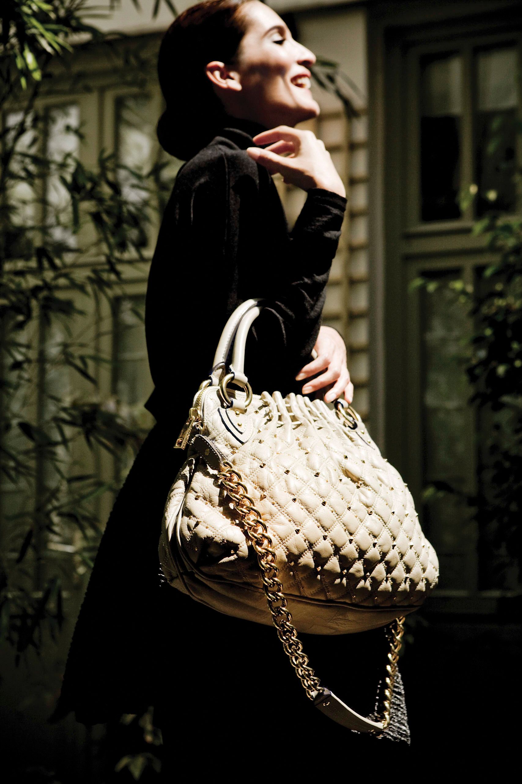 Woman in black dress smiling while holding a cream quilted handbag with gold chain strap outdoors.