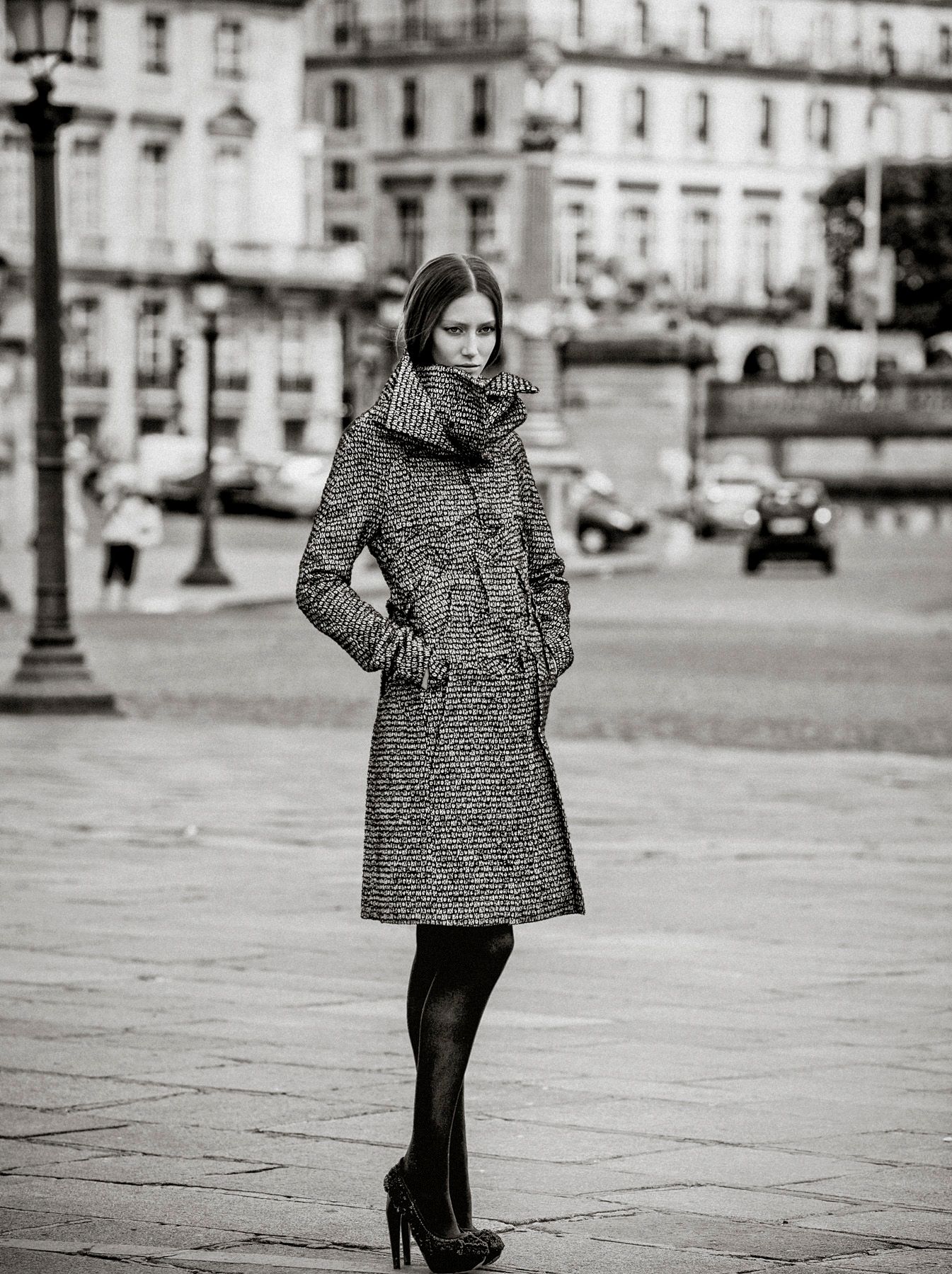 A stylish woman in a tweed coat and heels poses on a cobblestone plaza in a black and white urban photo.