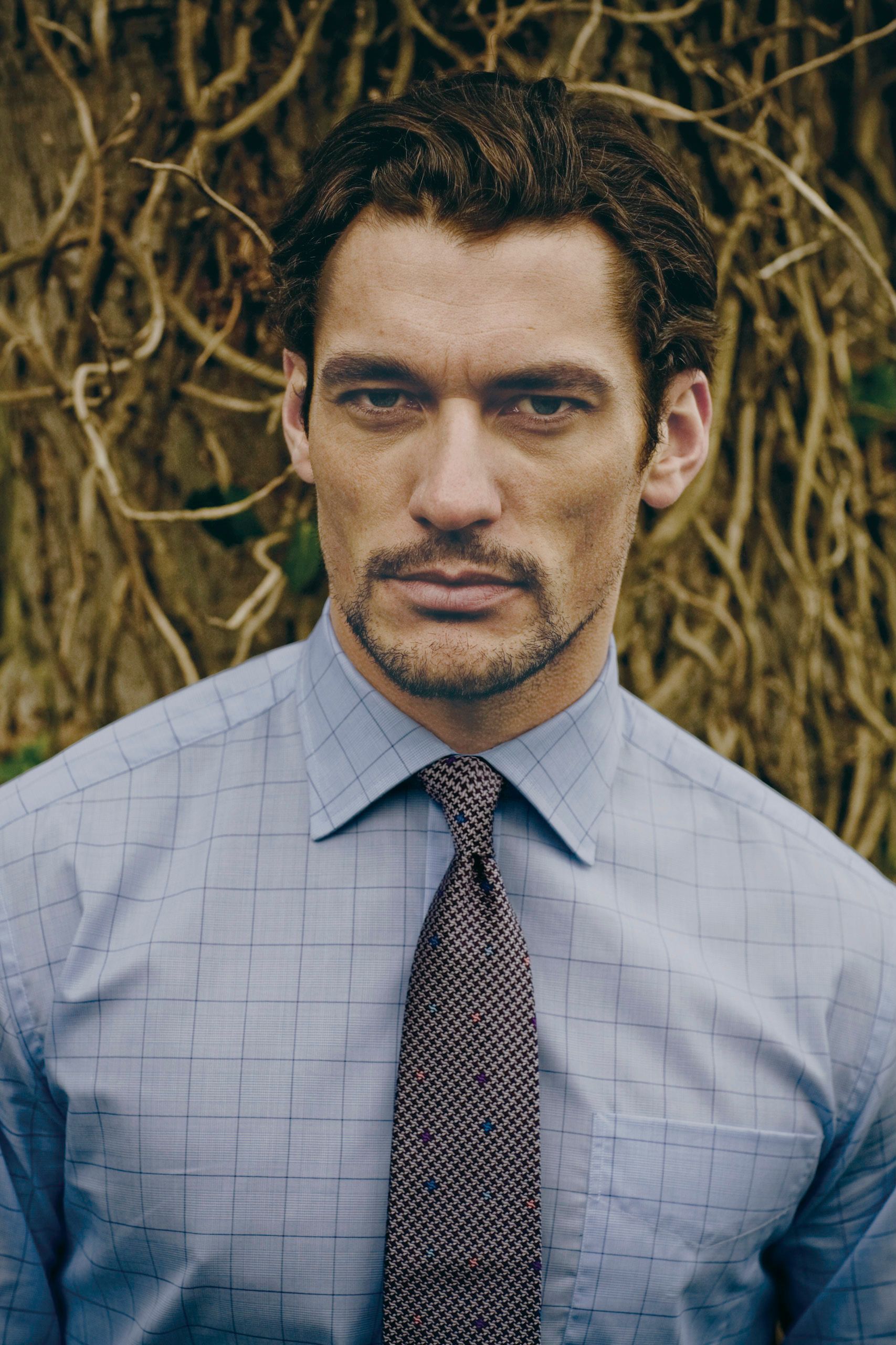 Man in blue checkered shirt and brown tie with short beard stands before ivy-covered wall.