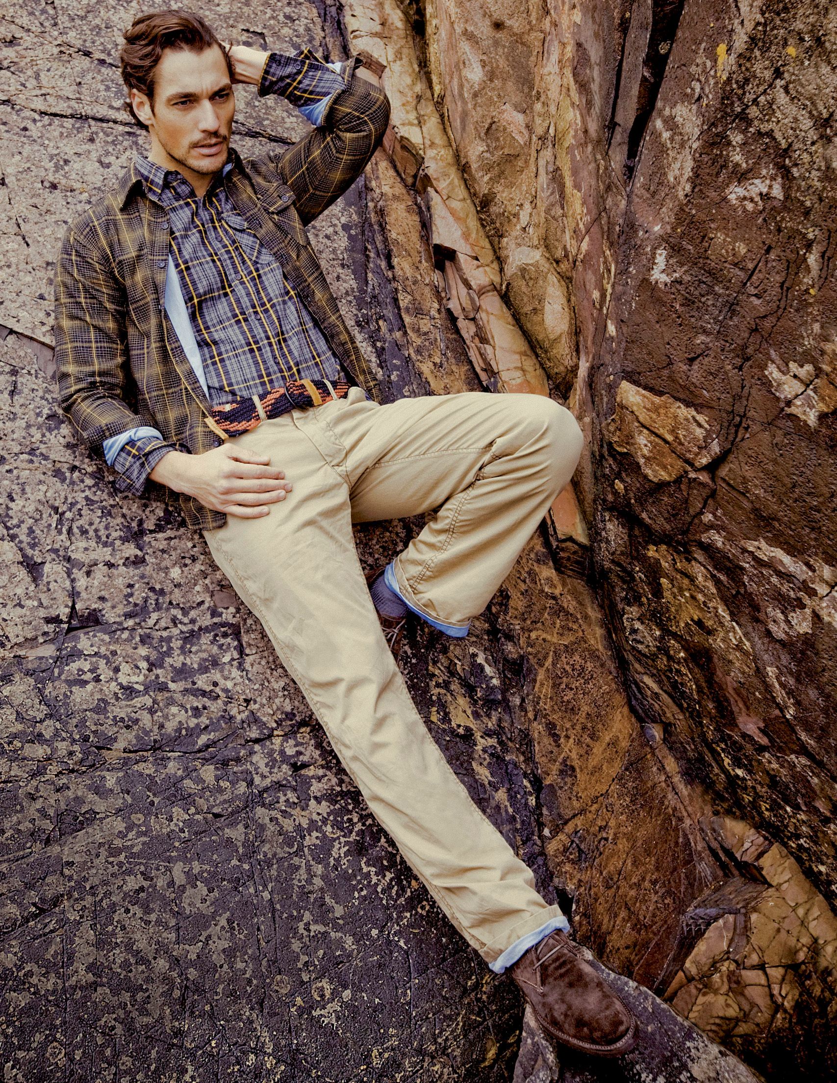 Man in plaid shirt and khaki pants leaning against rocky terrain outdoors.