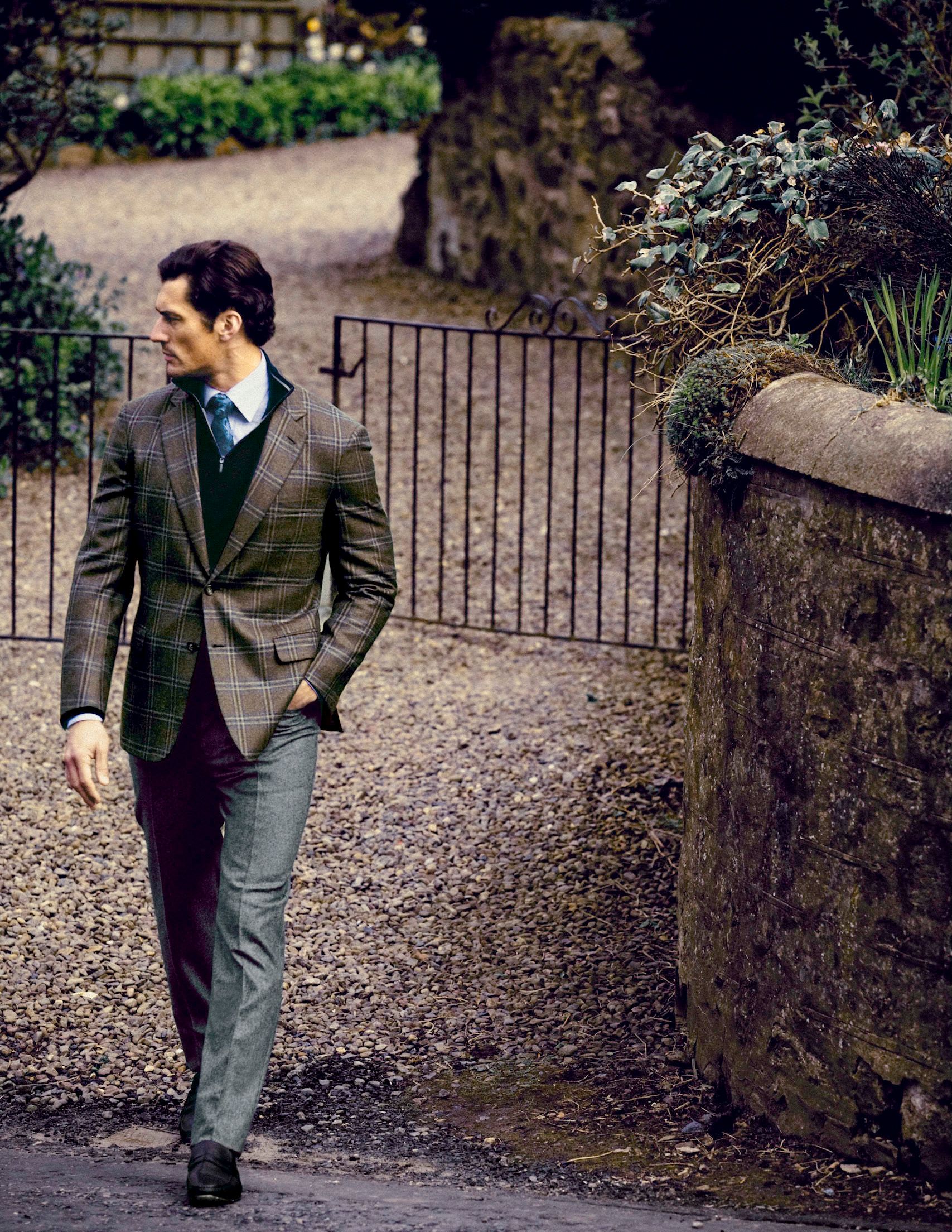 A well-dressed man in a plaid blazer and grey trousers walks along a gravel path beside a stone wall.