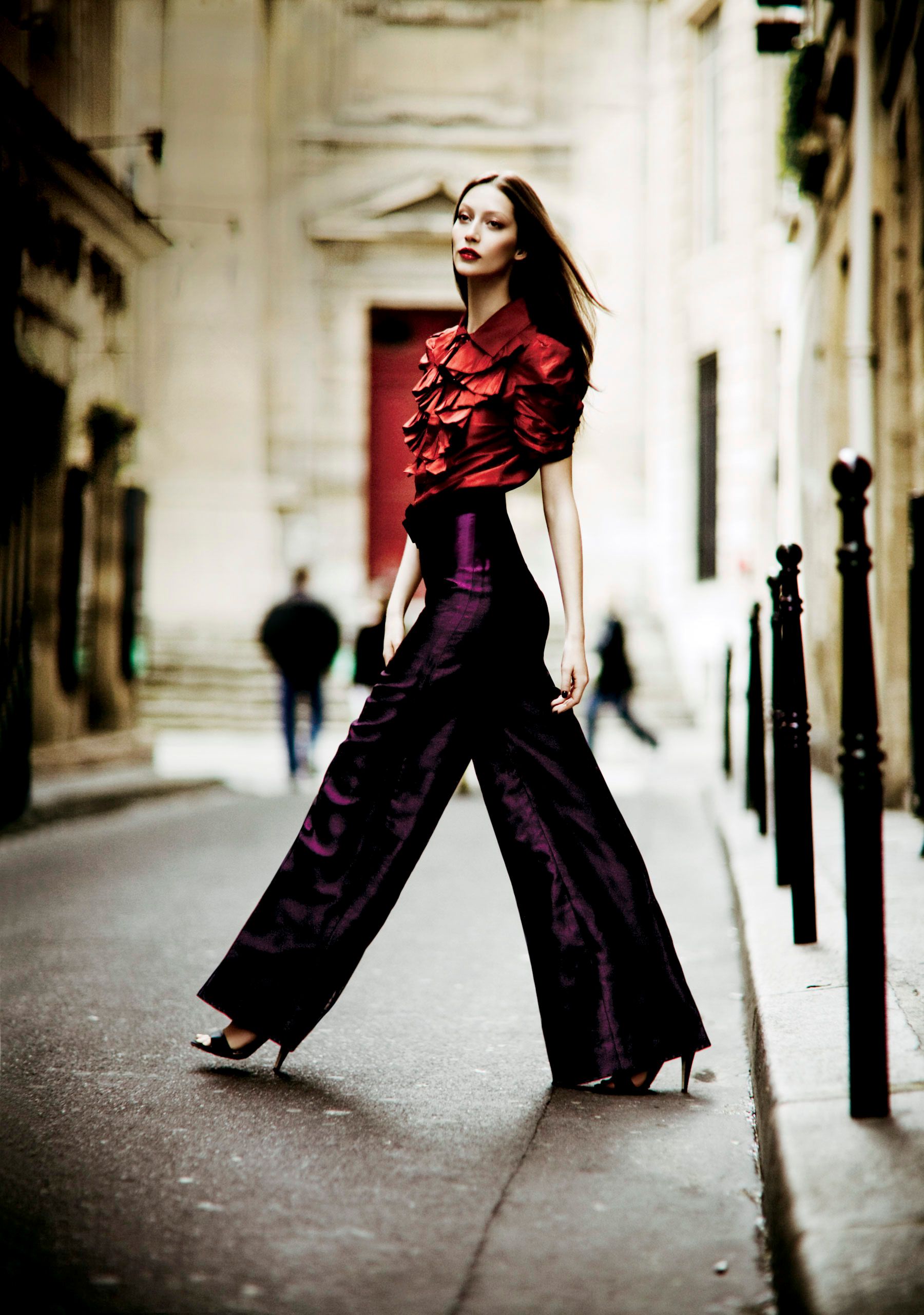 A model poses on a Parisian street in a red ruffled top and wide-leg purple satin trousers with black heels.