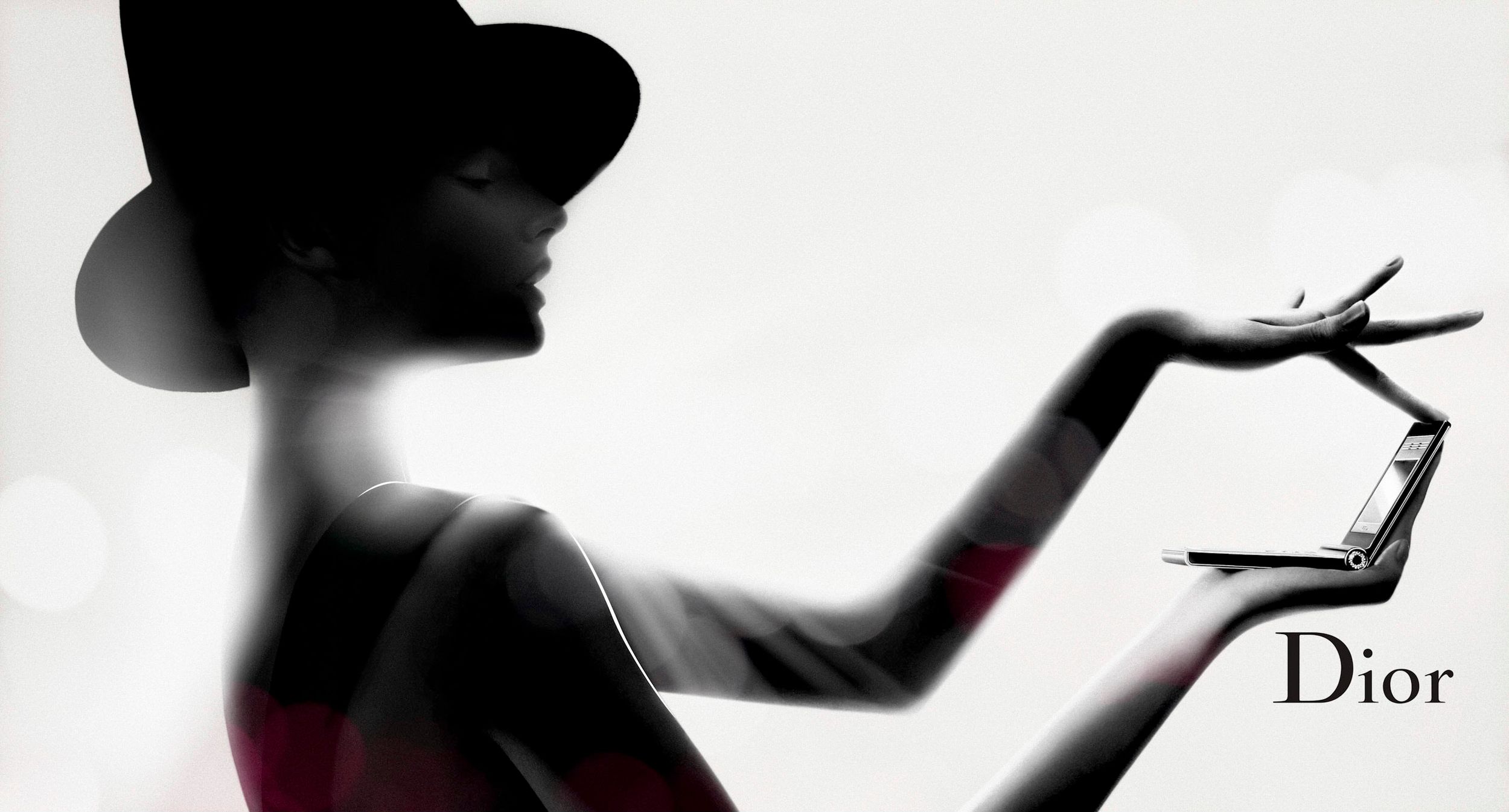Silhouette of elegant woman in wide-brim hat holding Dior cosmetics against soft bokeh background in luxury beauty ad.