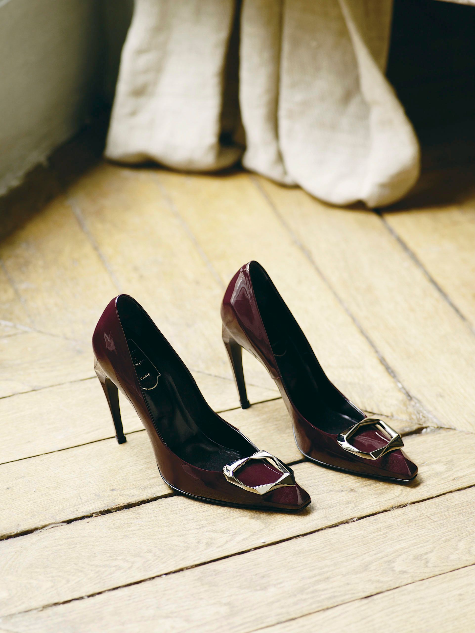 Burgundy patent leather pointed-toe stiletto heels with silver buckle detail on wooden floor.