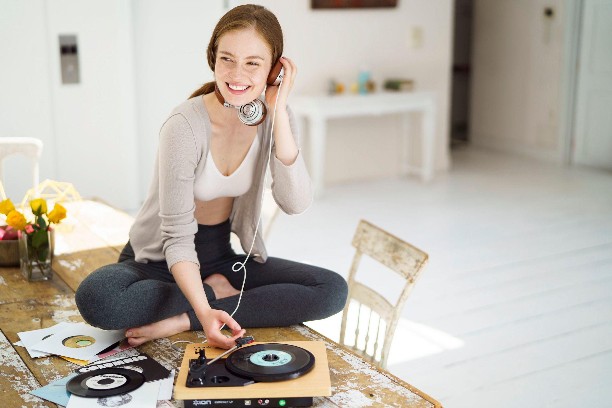 Young woman sitting cross-legged on floor, smiling while listening to vinyl record on retro turntable with headphones.