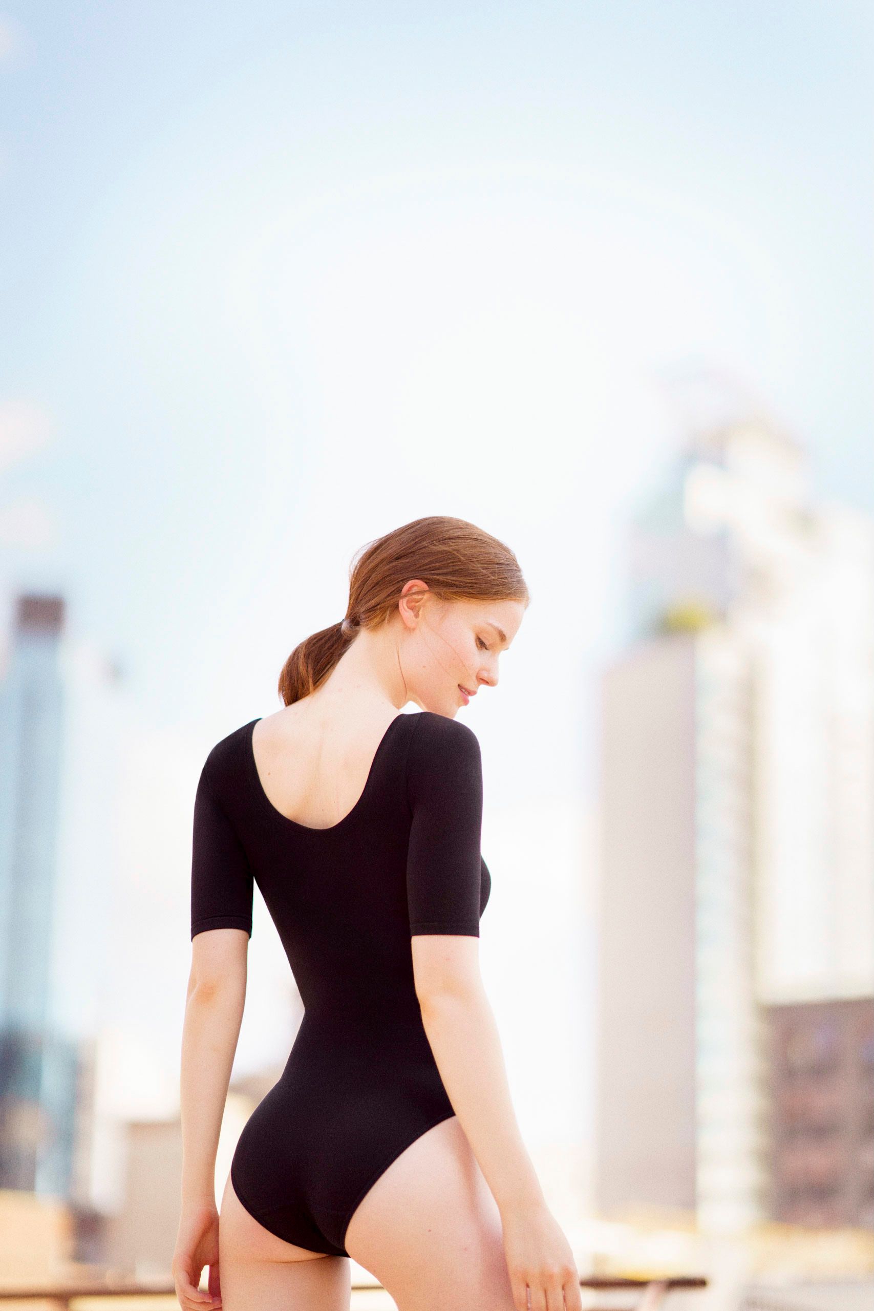 Young woman in black leotard poses on rooftop with city skyline in soft focus background.