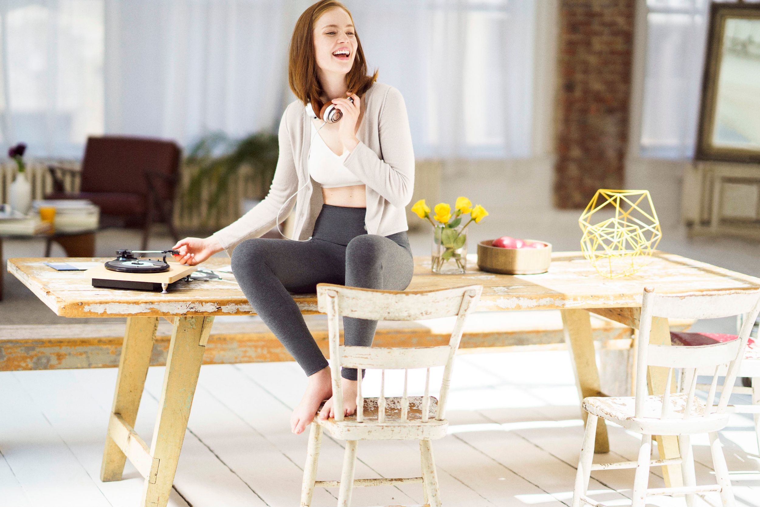 Young woman sitting on wooden table, wearing headphones, smiling in bright modern living room with vinyl record player.