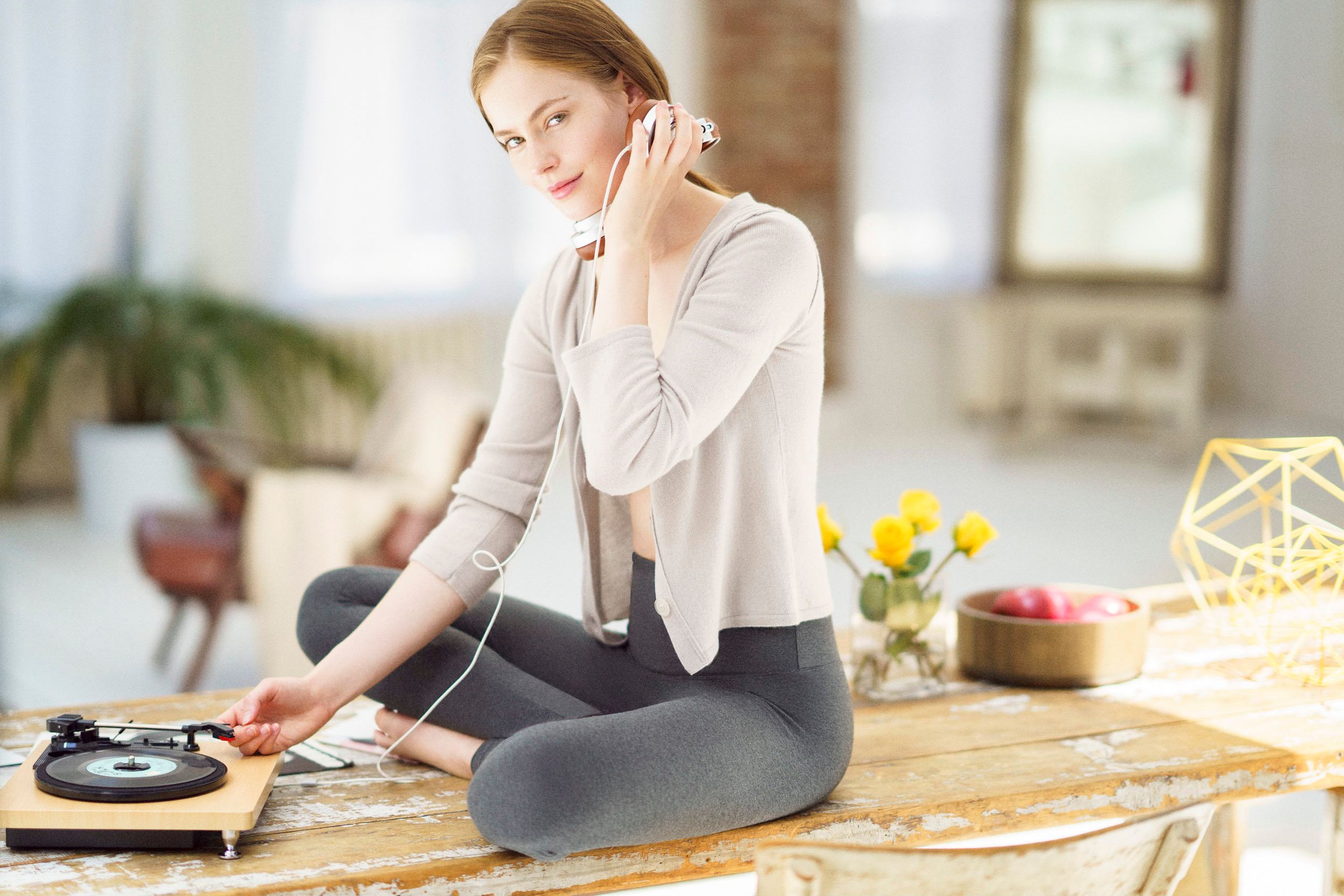 Young woman sitting cross-legged on table, listening to music via headphones connected to a vinyl record player.
