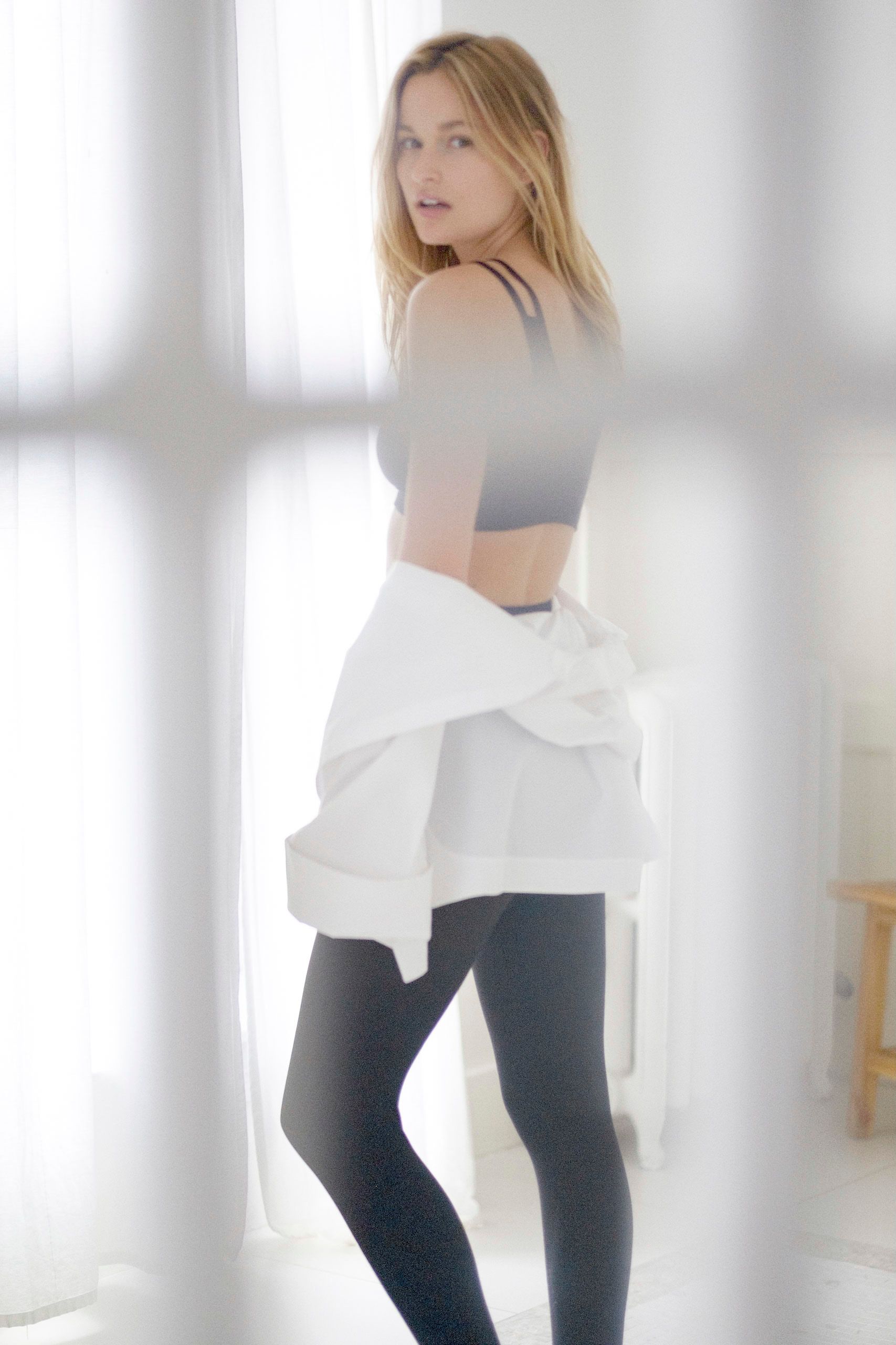 Woman in gray sports bra and black leggings holding white shirt near bright window.