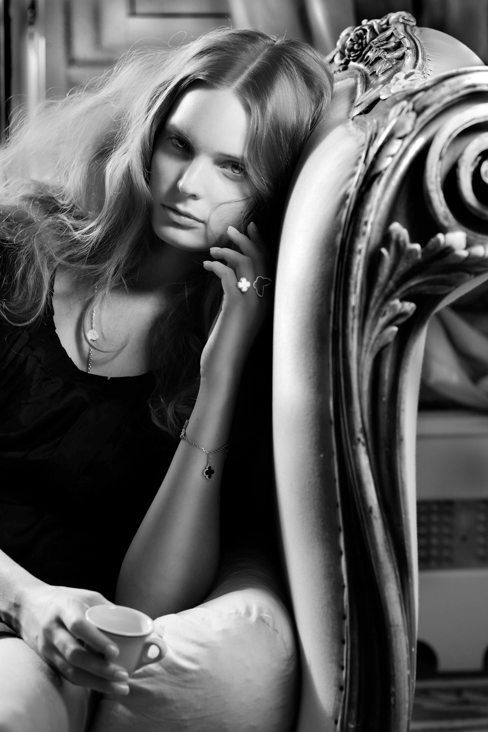 Elegant woman in black top holding a teacup, seated in ornate chair, black and white fashion portrait.