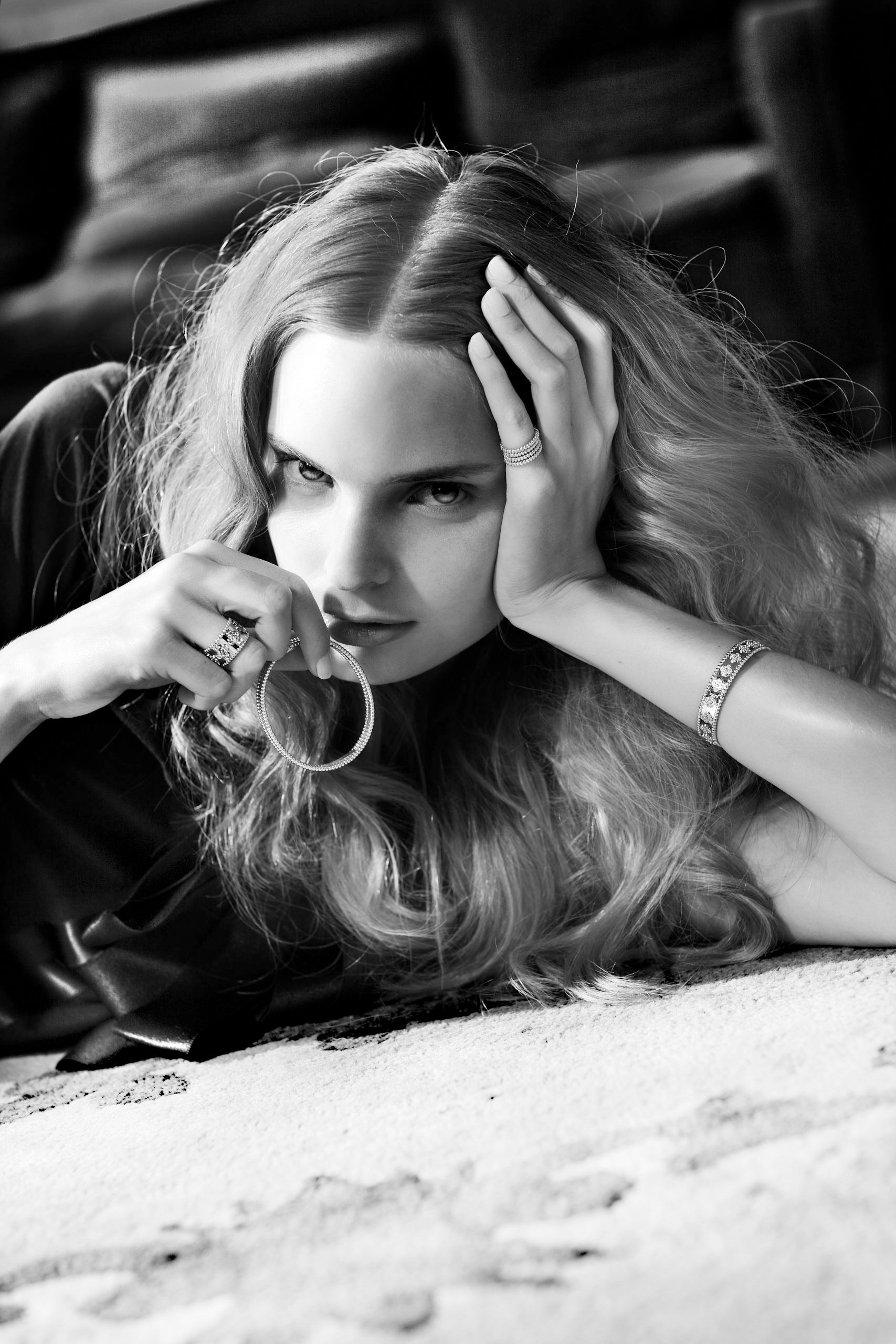 Black and white photo of a woman with long wavy hair lying down, wearing jewelry, rings and bracelet, looking at camera.