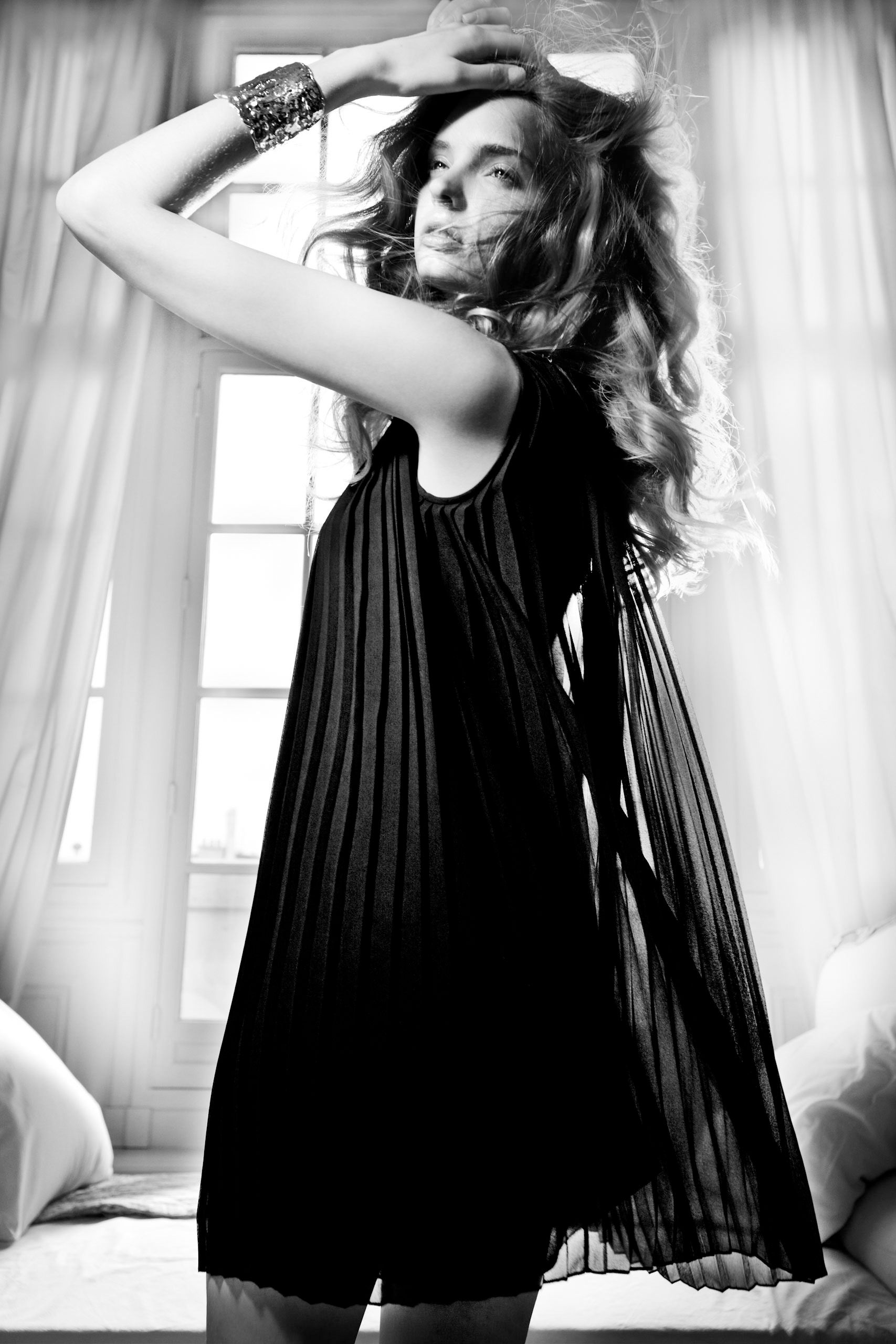 Black and white photo of a woman in a flowing pleated black dress posing elegantly near a bright window.