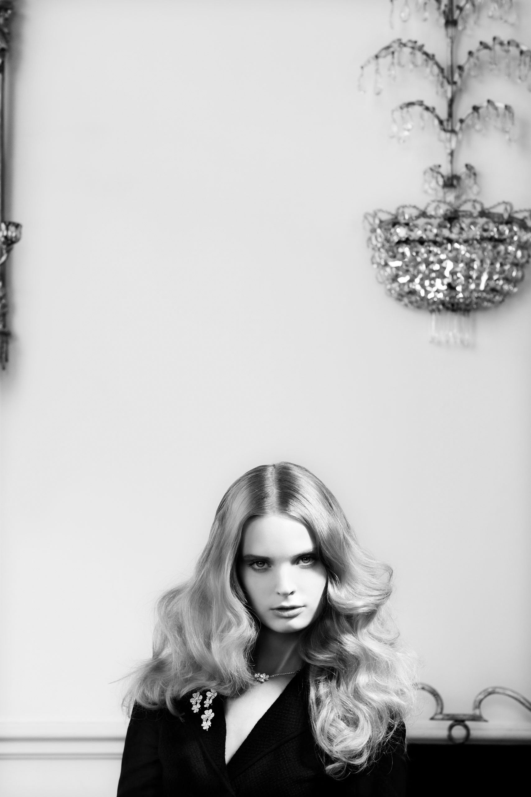 Black and white fashion portrait of a woman with long wavy blonde hair wearing a dark blazer with brooch by chandelier.