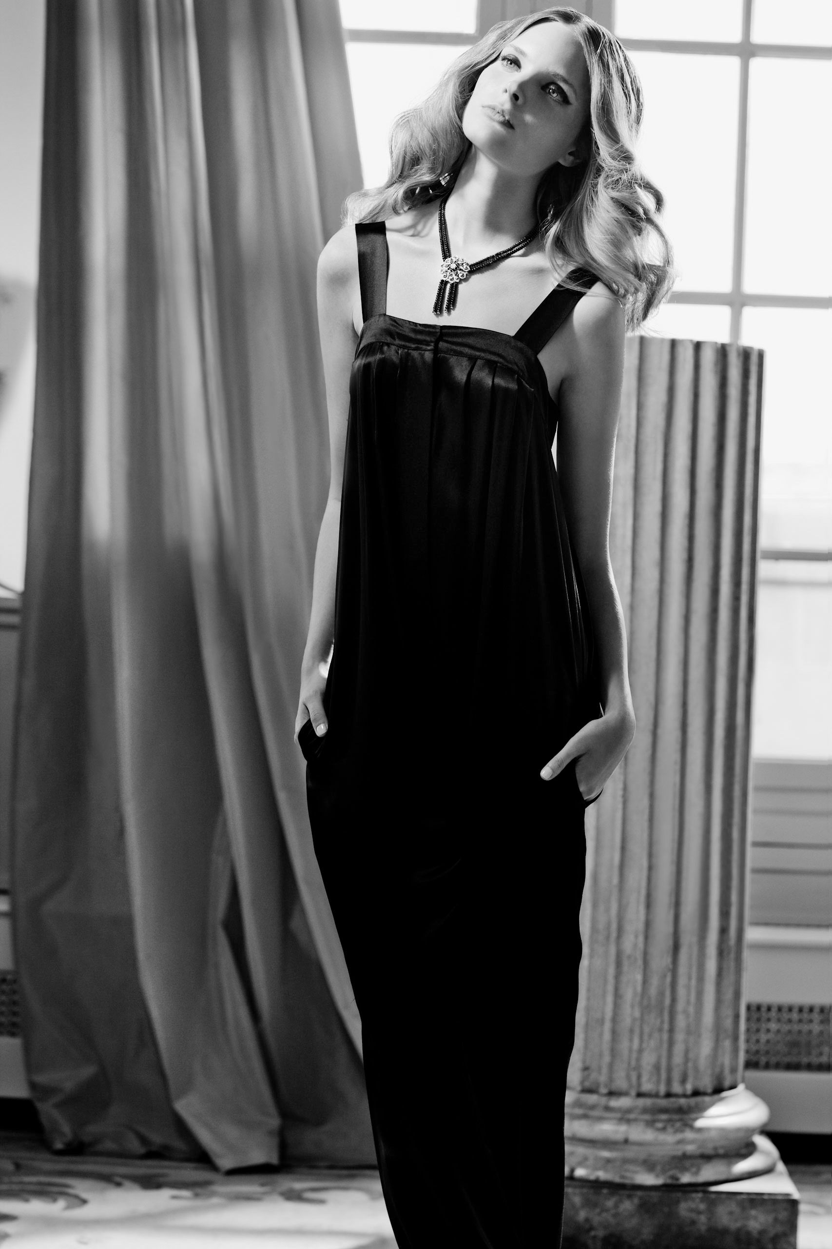 Black and white photo of a blonde woman in a long black dress with a pendant necklace posing by curtains.