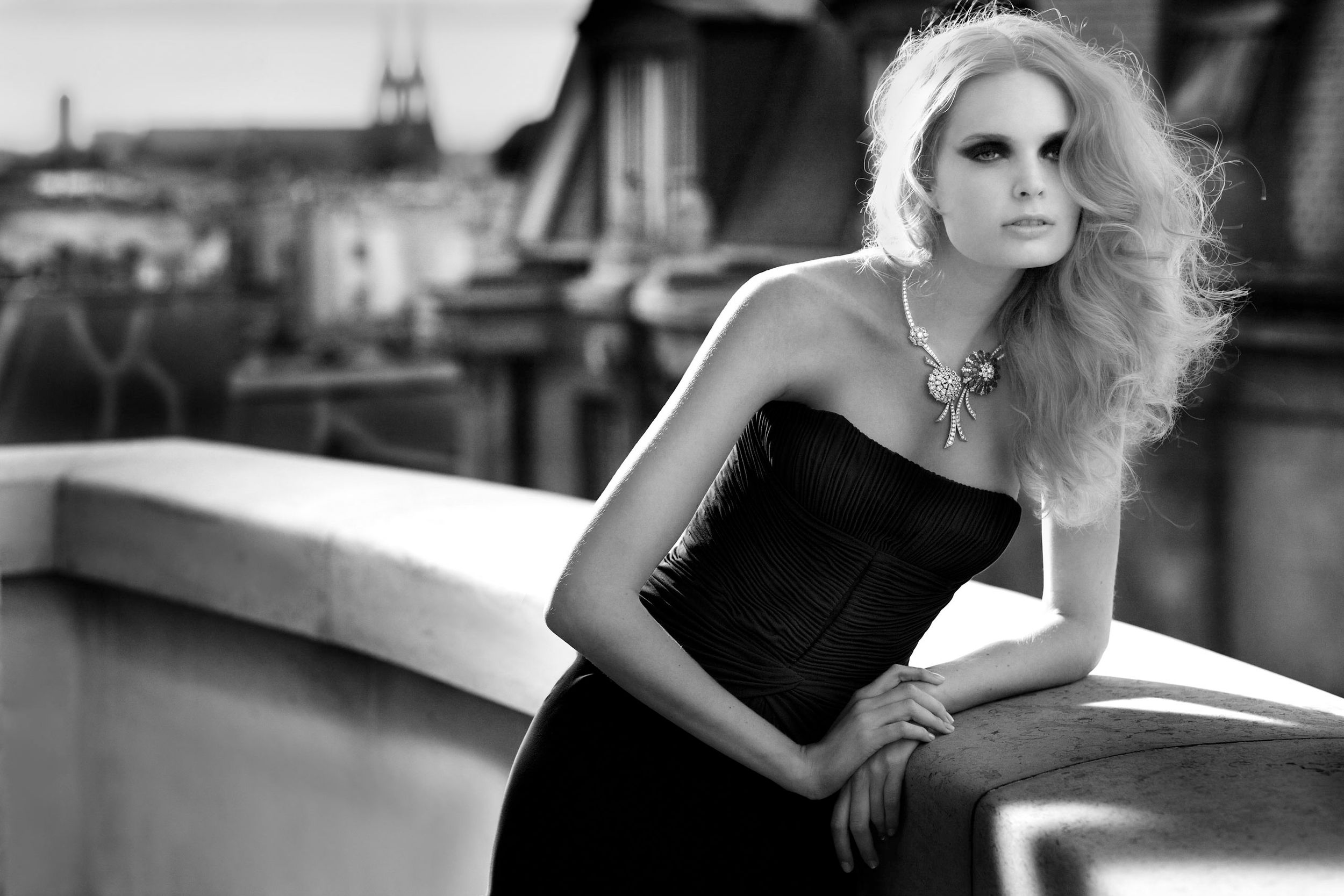 Elegant blonde woman in black strapless dress posing on rooftop terrace with city skyline in black and white.
