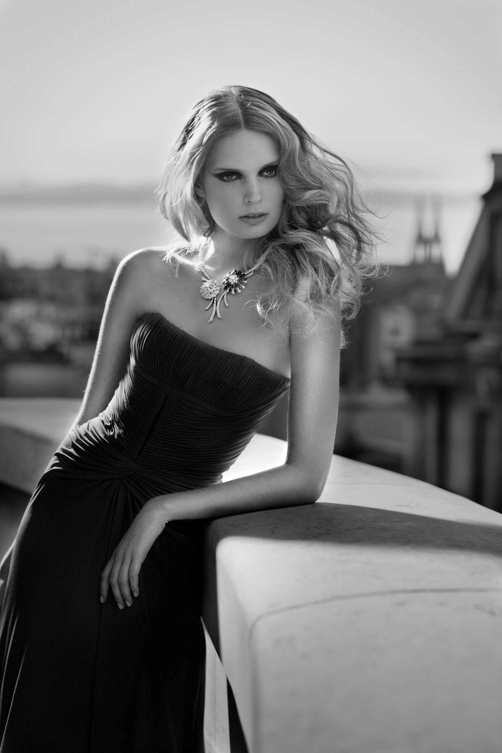 Elegant blonde woman in strapless black gown with statement necklace posing on rooftop in black and white photo.