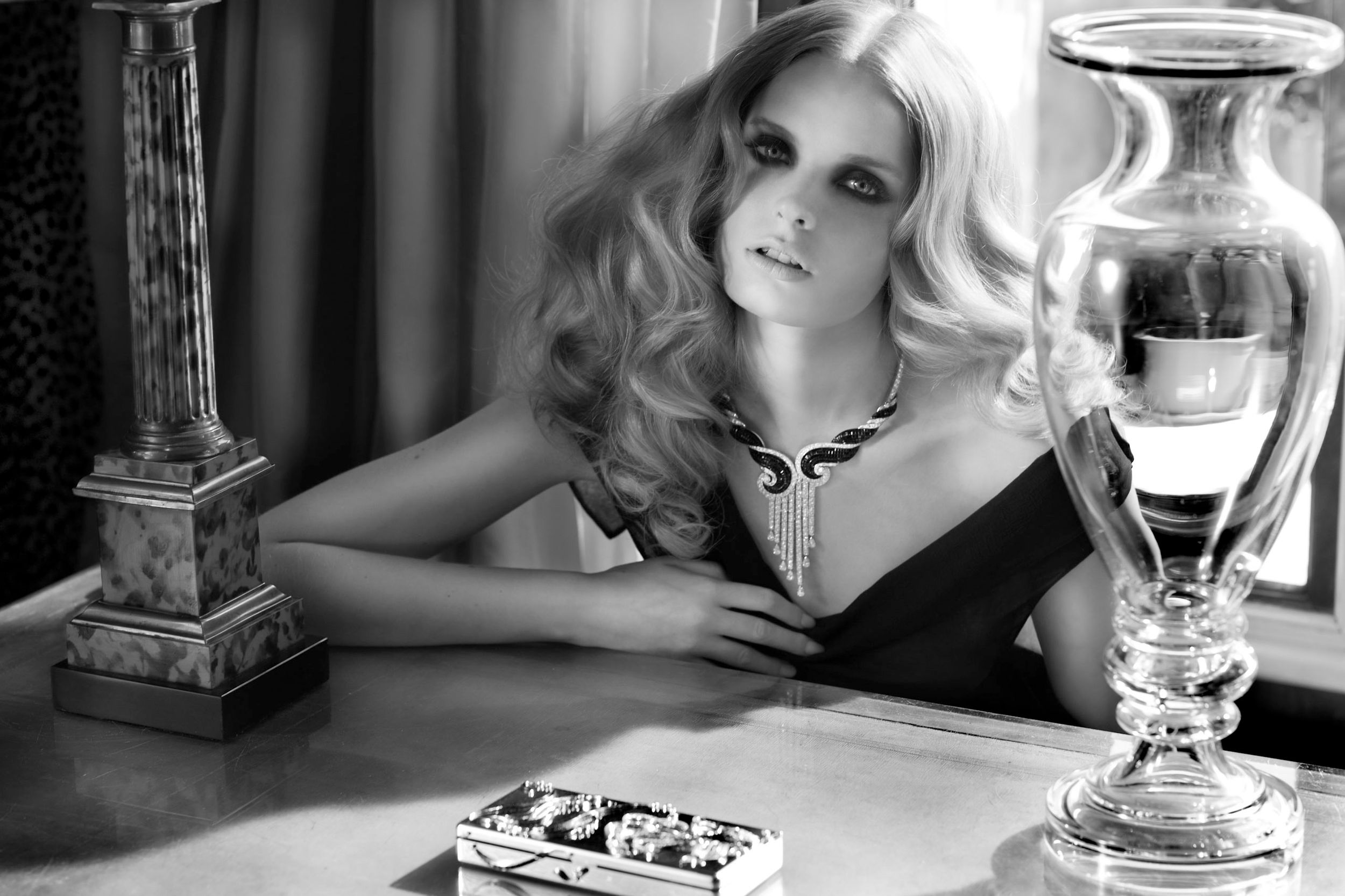 Elegant blonde woman in black dress with necklace poses at table with vase in black and white photo.