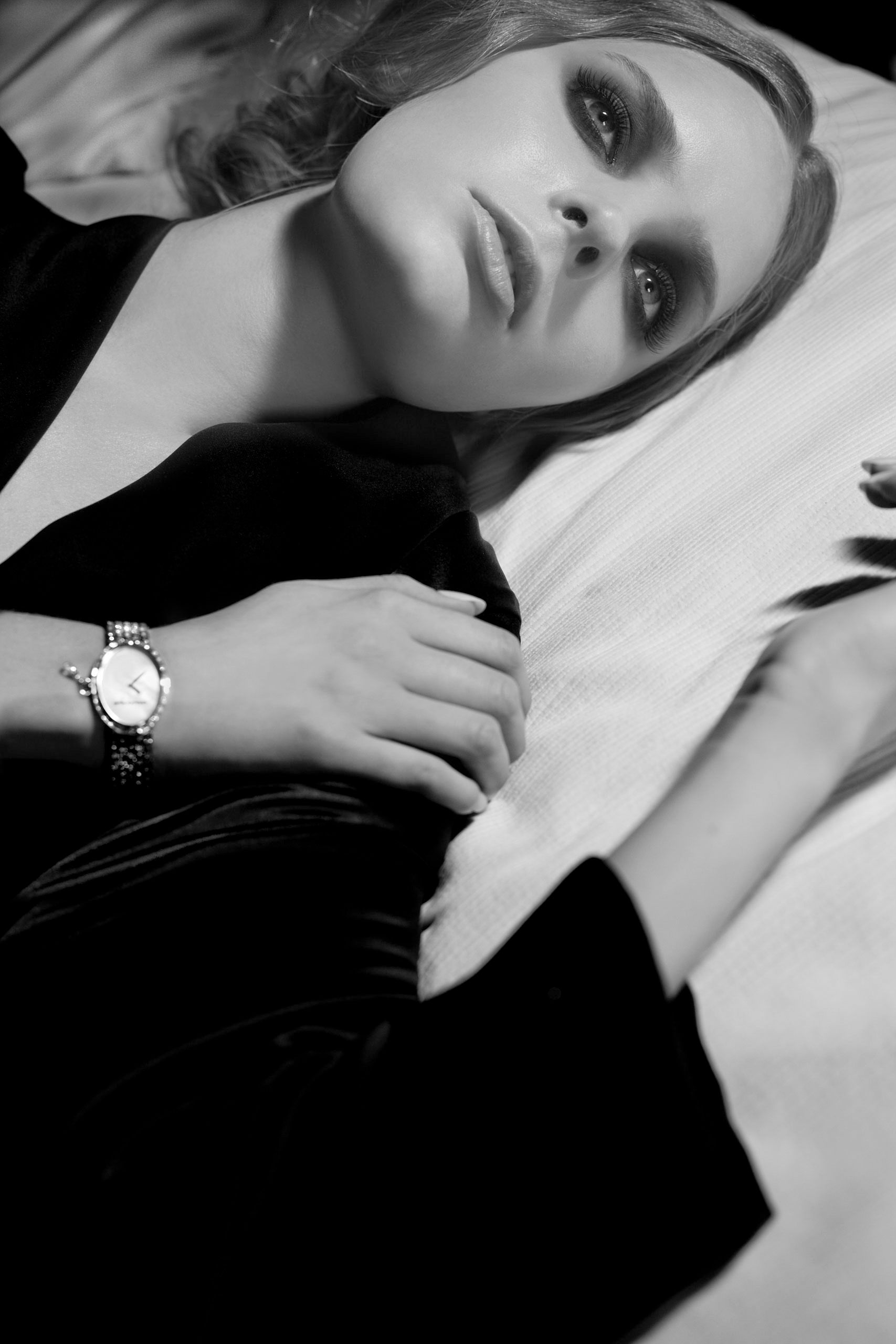 Black and white photo of a woman in velvet blazer wearing a diamond watch, lying elegantly against white fabric.