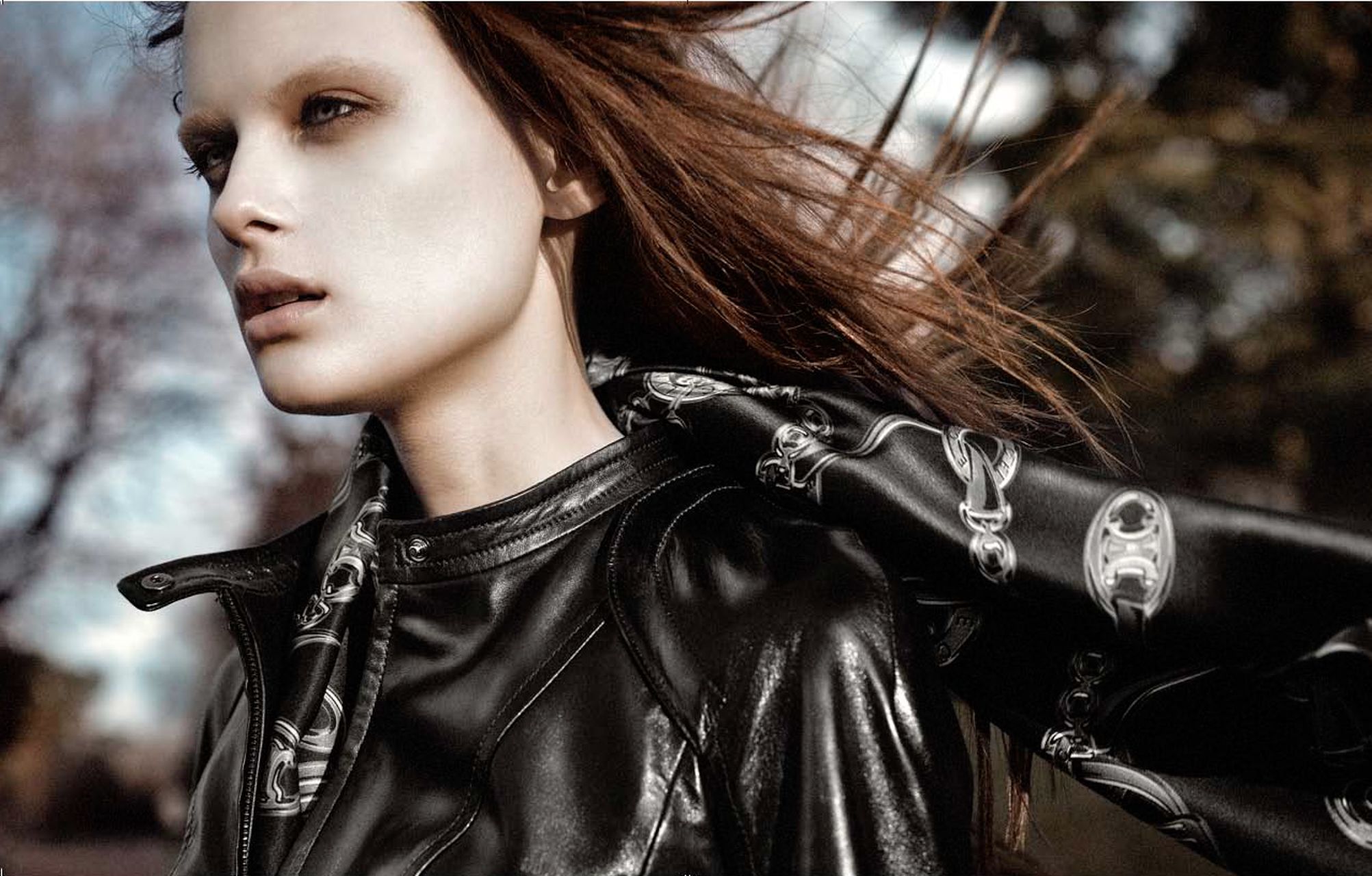 Model in black leather jacket with silver hardware poses outdoors with windswept hair in editorial fashion shoot.