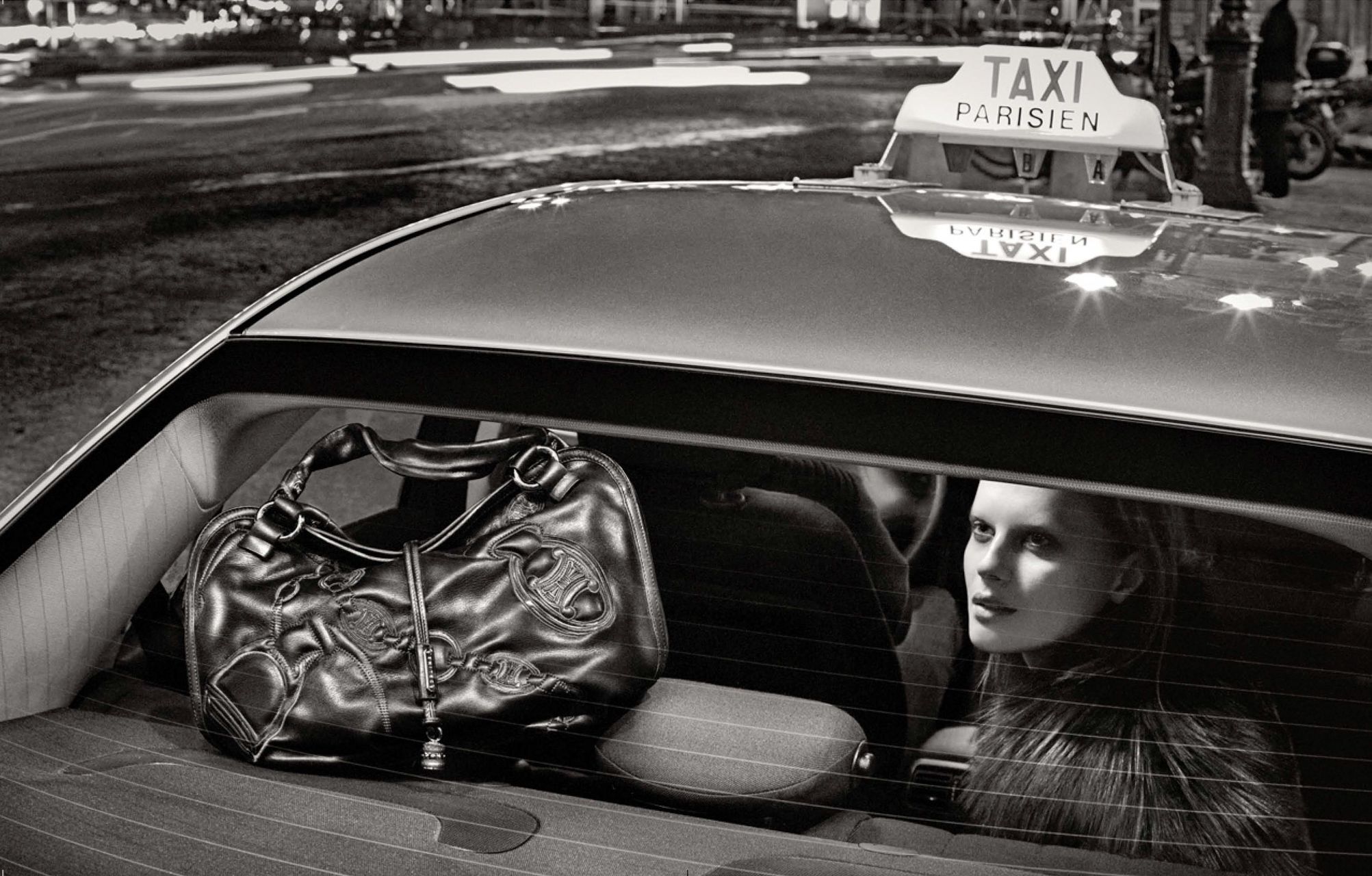 Black and white photo of a woman sitting in a Parisian taxi cab with a large handbag on the seat beside her.