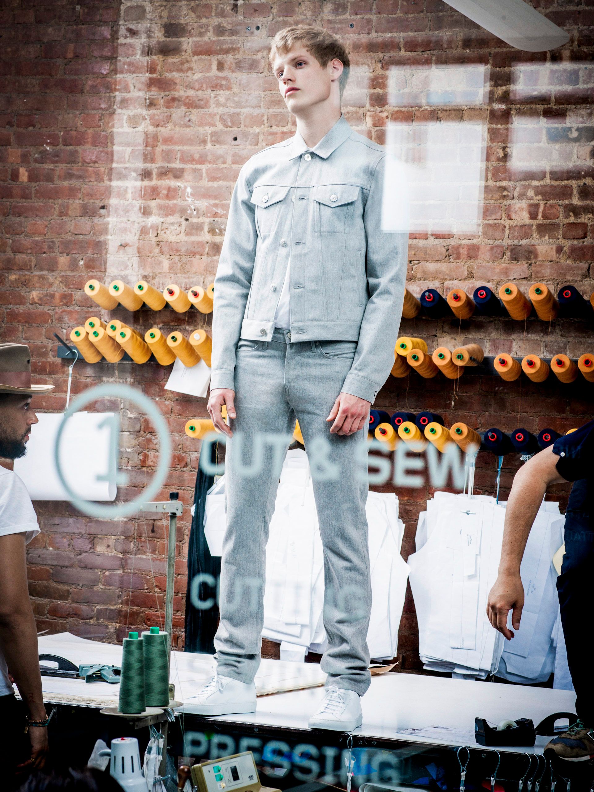 A male model stands on a table wearing a light gray denim jacket and matching pants at a fashion show near a brick wall.