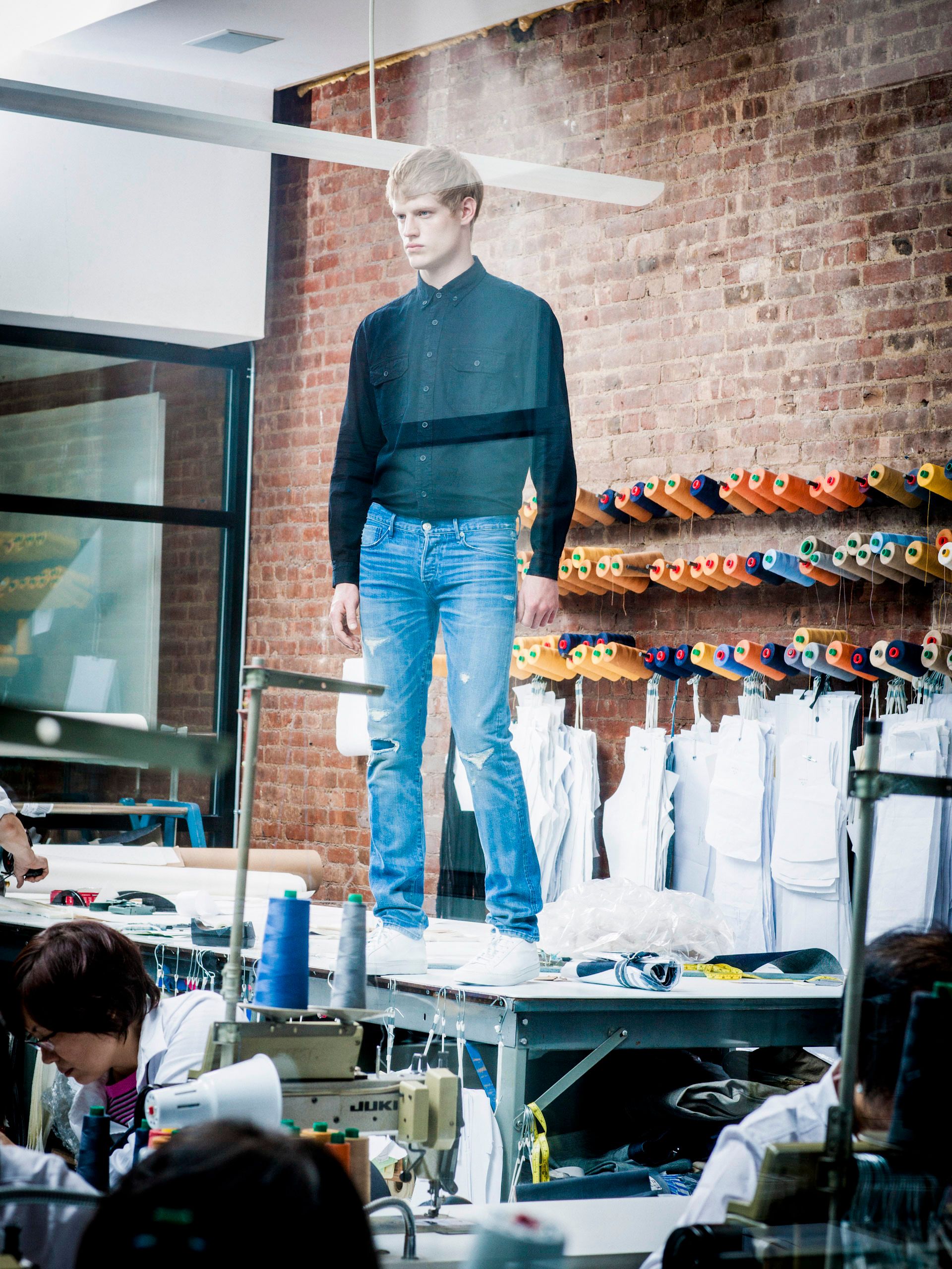 Male model in black shirt and blue jeans stands on a worktable in a fashion design studio with brick walls.