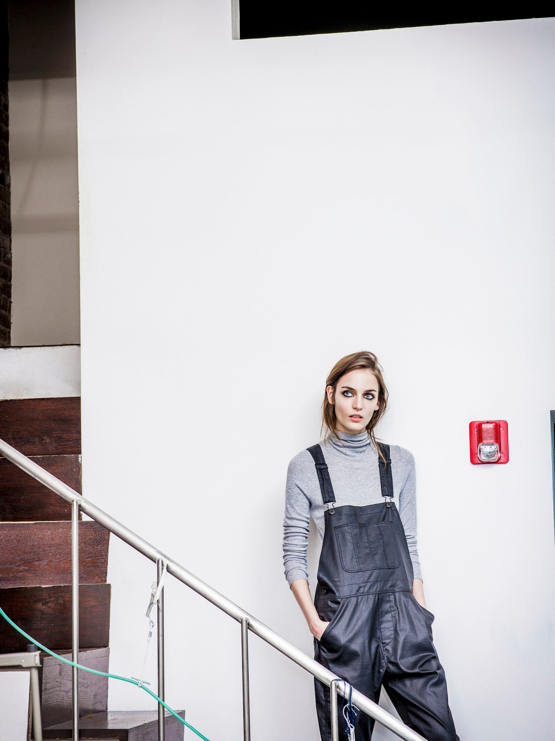 Woman in gray turtleneck and dark overalls leaning against white wall on staircase landing.
