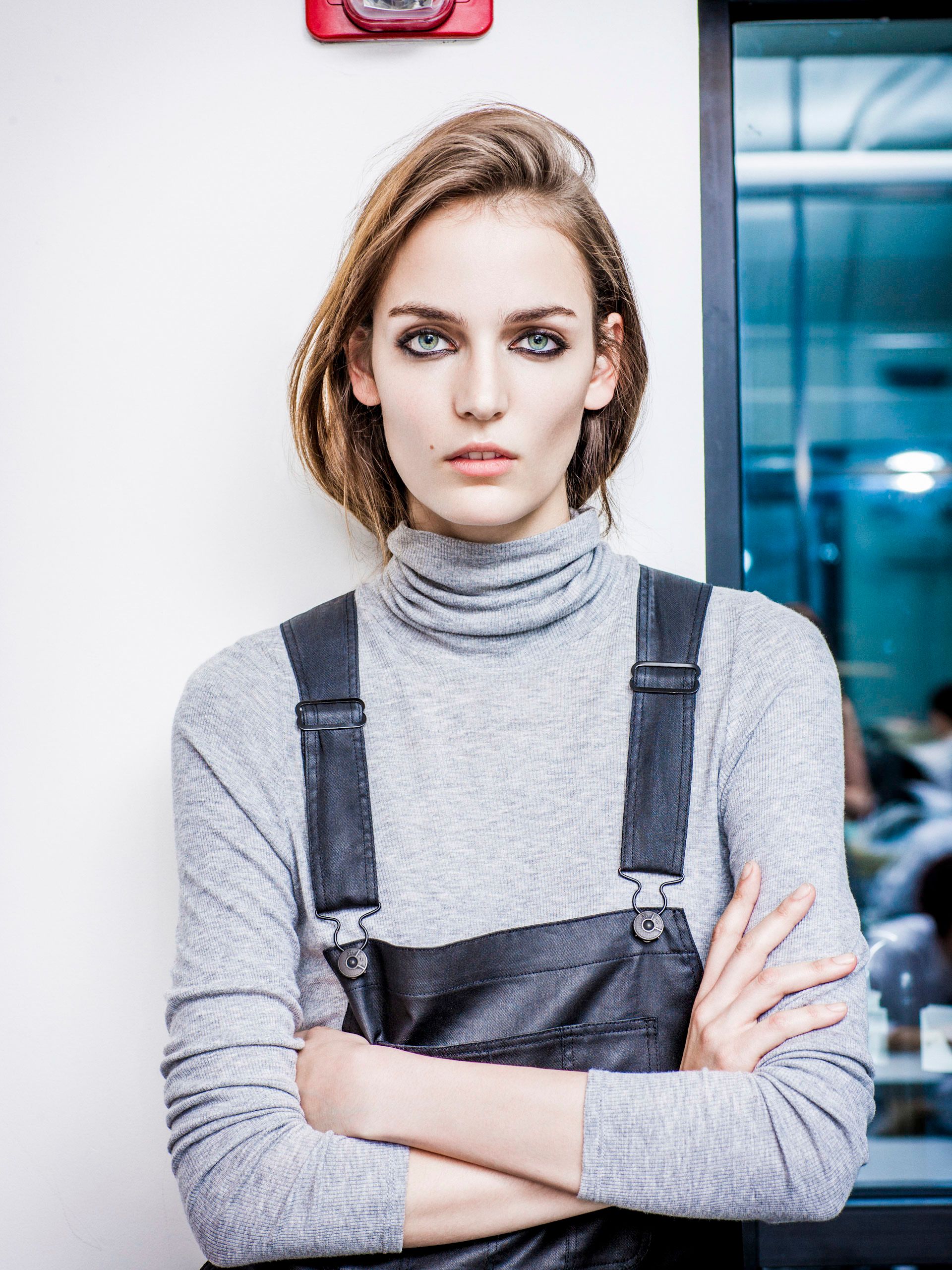 Woman in gray turtleneck and black overalls stands with arms crossed against a white wall.