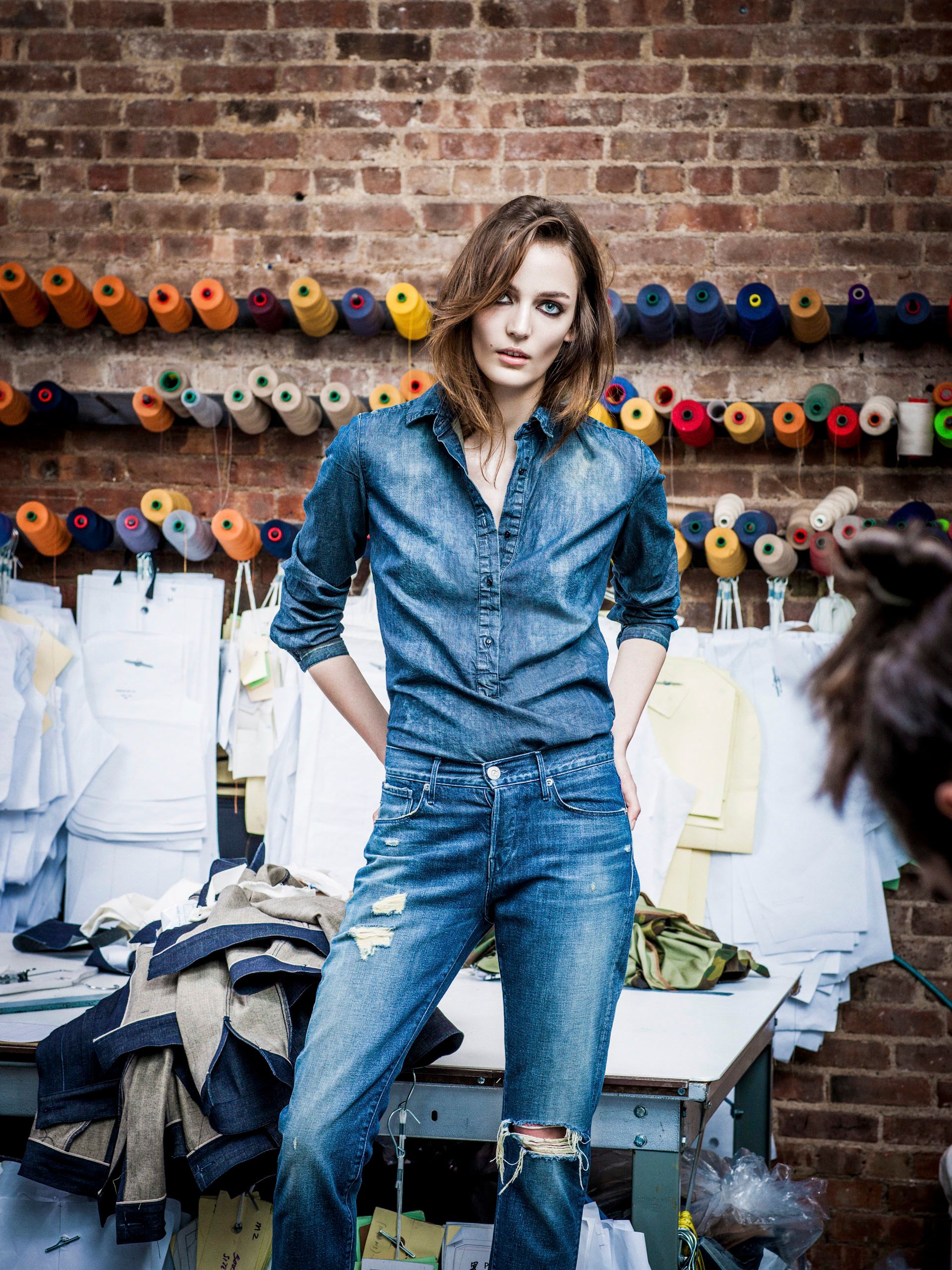 Woman in denim shirt and jeans poses in a textile workshop with colorful thread spools on brick wall.