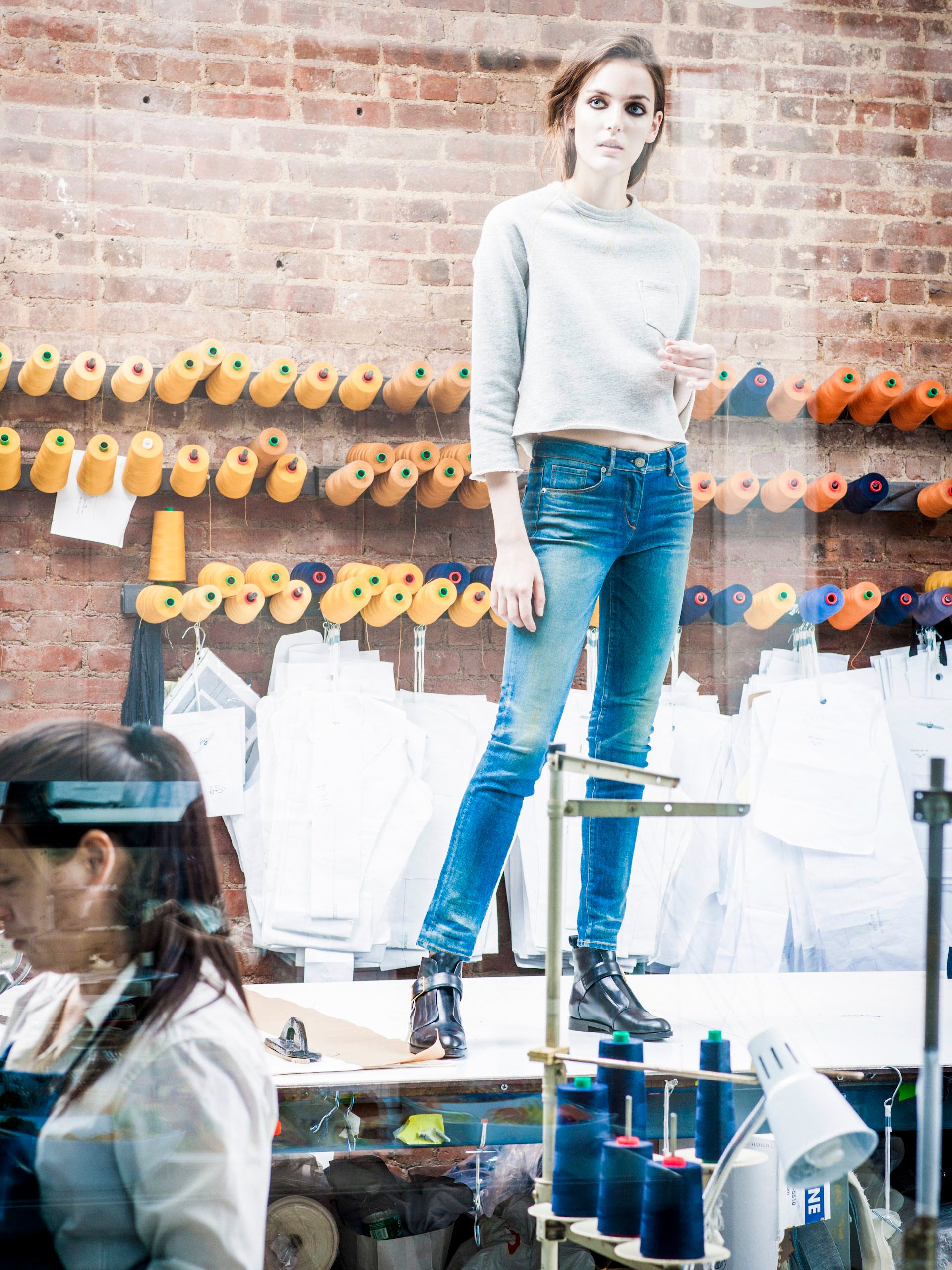 A model stands on a table in a denim studio, wearing blue jeans and a gray sweatshirt, surrounded by colorful thread spools.