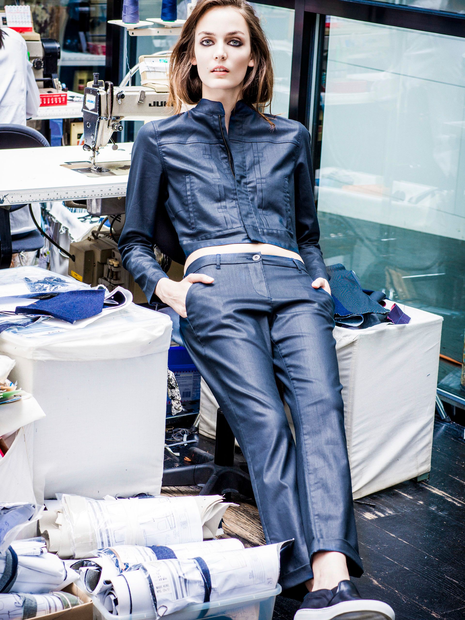 Woman in navy blue jumpsuit poses among fabric rolls in a tailoring workshop environment.