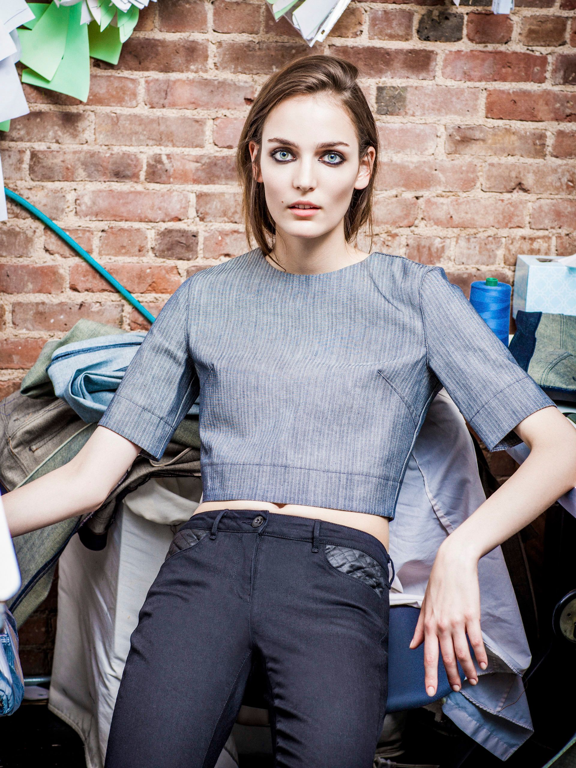 Woman in gray crop top and black pants poses against brick wall in a fashion studio setting.