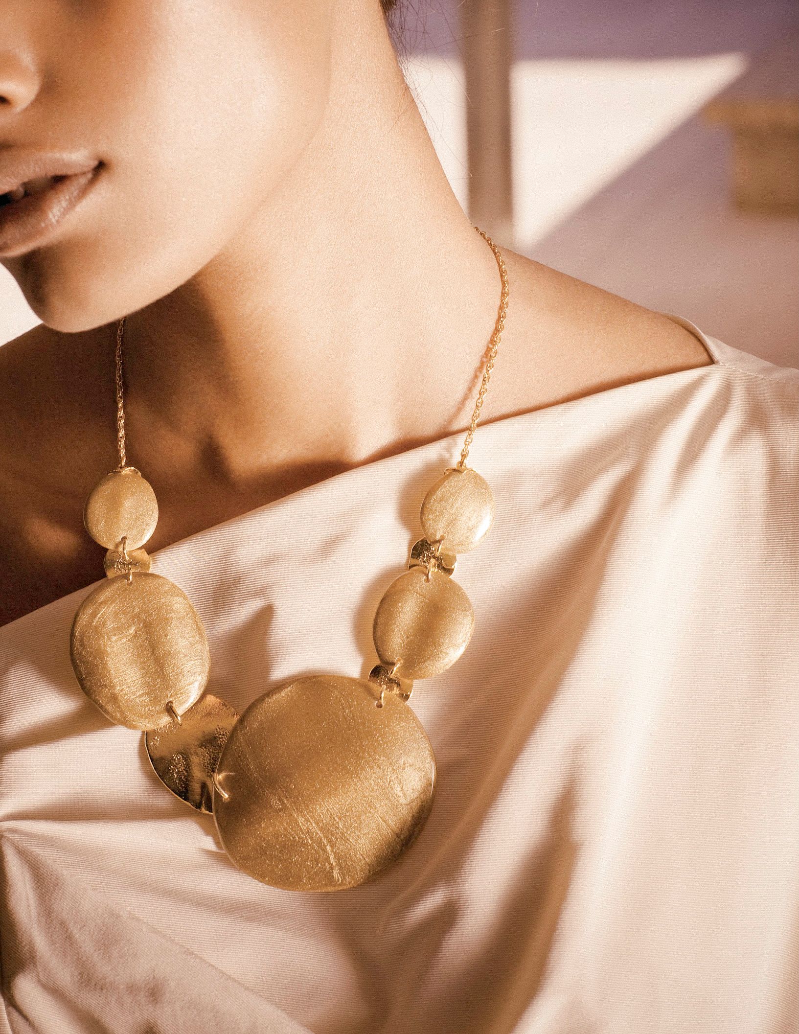 Gold statement necklace with large oval and round brushed metal pendants on a delicate chain, worn with a cream silk top.