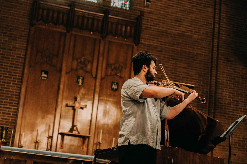 A male violinist performs in a historic brick church with wooden panels and a cross visible in the background.