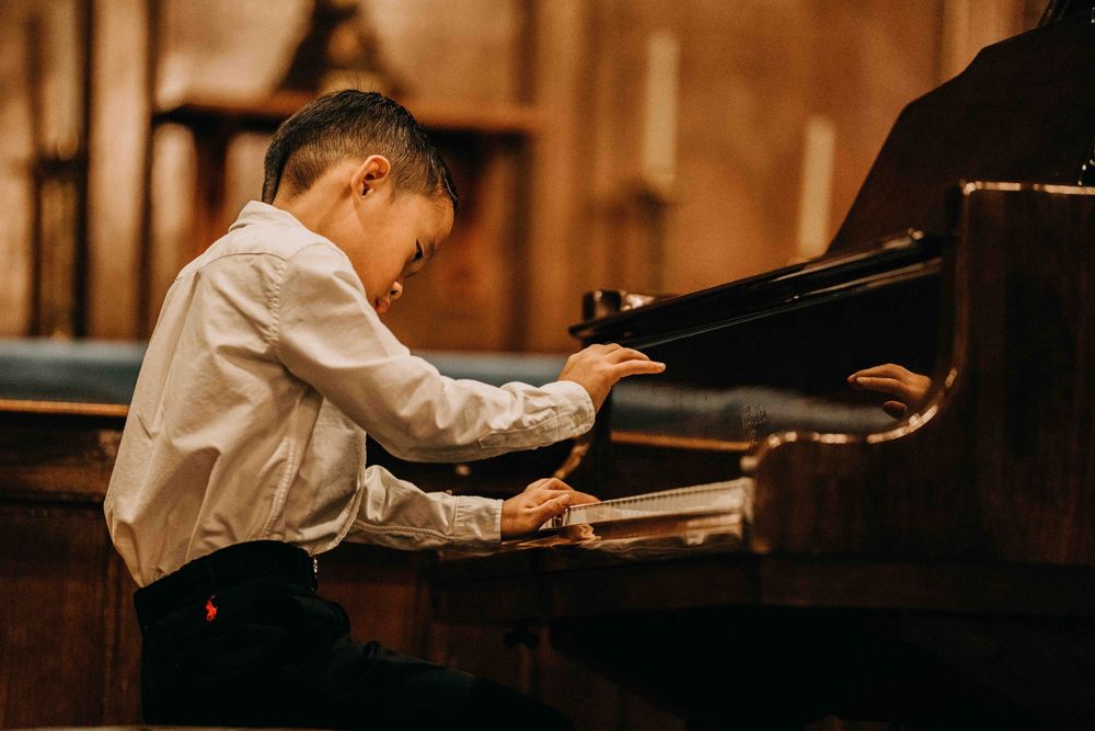 Young boy in white shirt playing grand piano during a recital in an elegant wood-paneled hall.