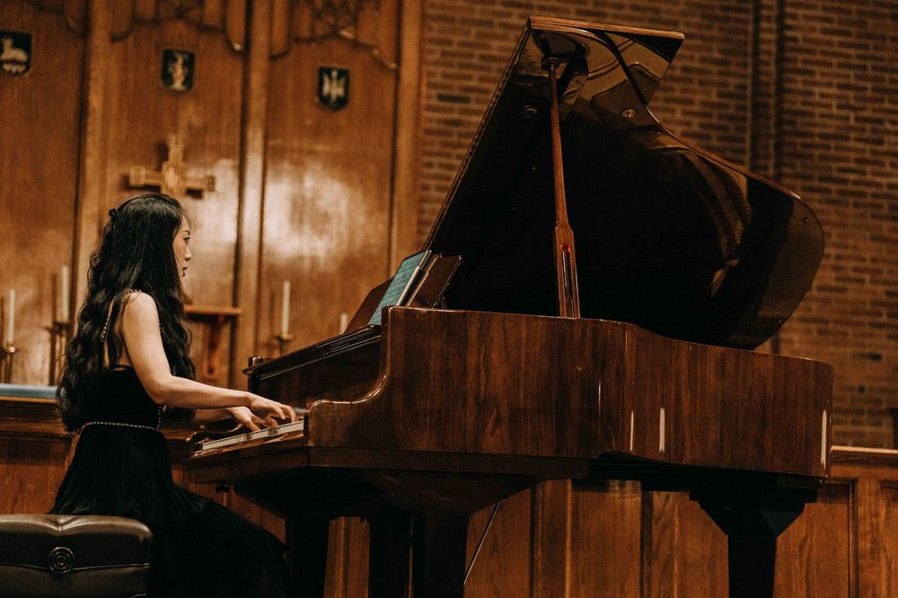A young woman plays a grand piano on stage in an elegant wood-paneled concert hall.