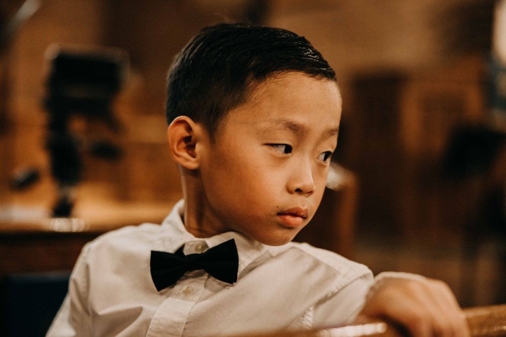 A young Asian boy in a white shirt and black bow tie sits thoughtfully in a church pew.