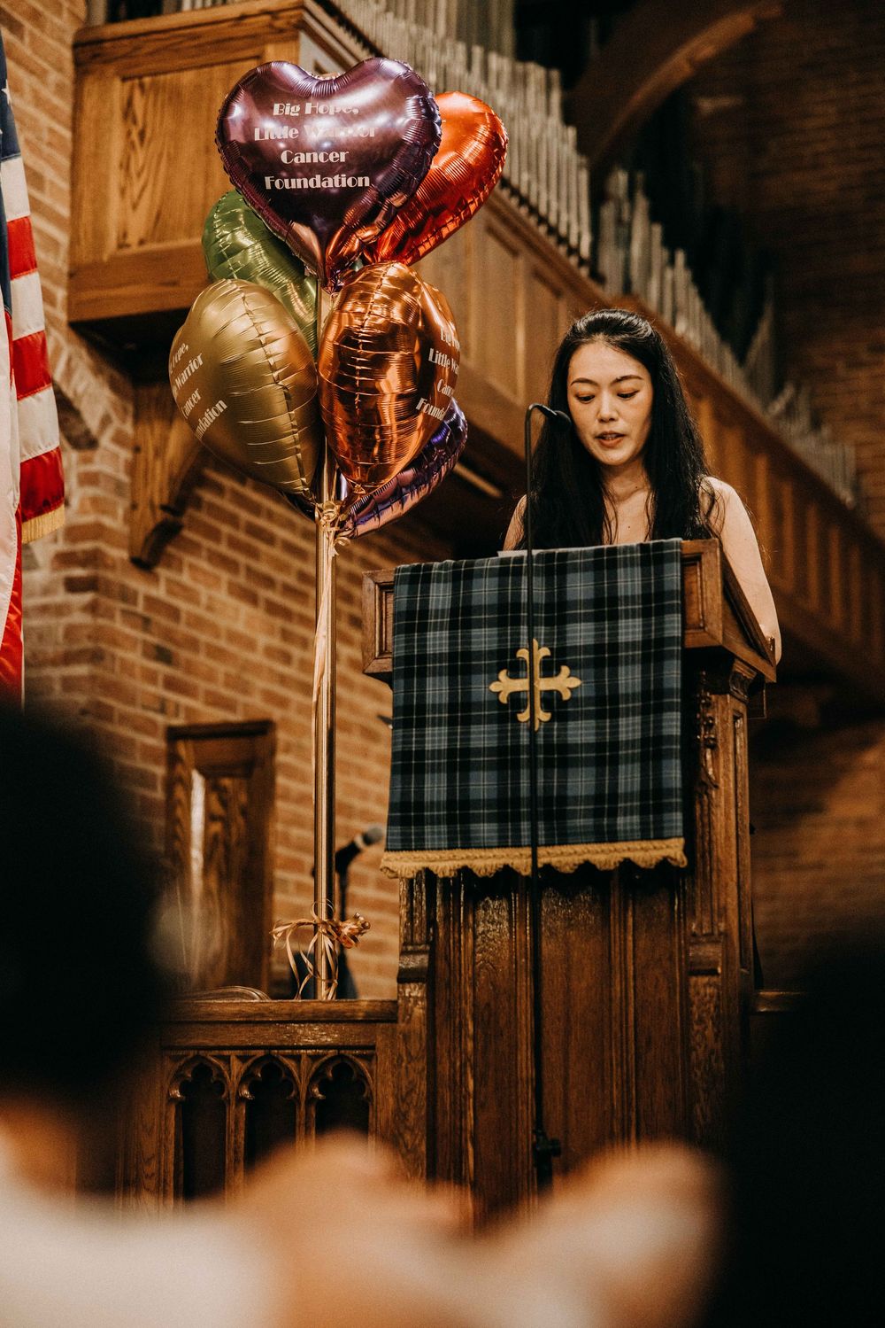 Woman speaking at a church podium with colorful heart-shaped balloons and a Cancer Foundation banner beside her.