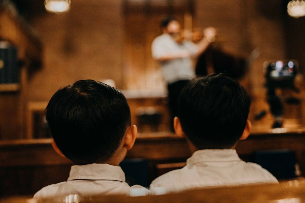 Two young boys in white shirts watch a violinist perform in a warmly lit venue.
