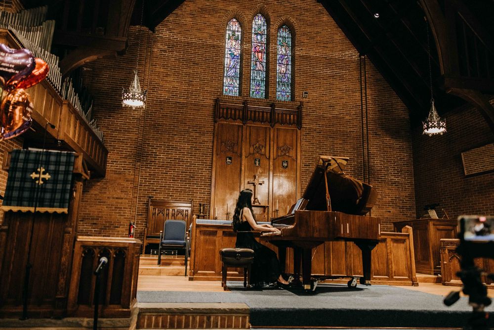 A pianist performs on a grand piano inside a stunning brick church with colorful stained glass windows.